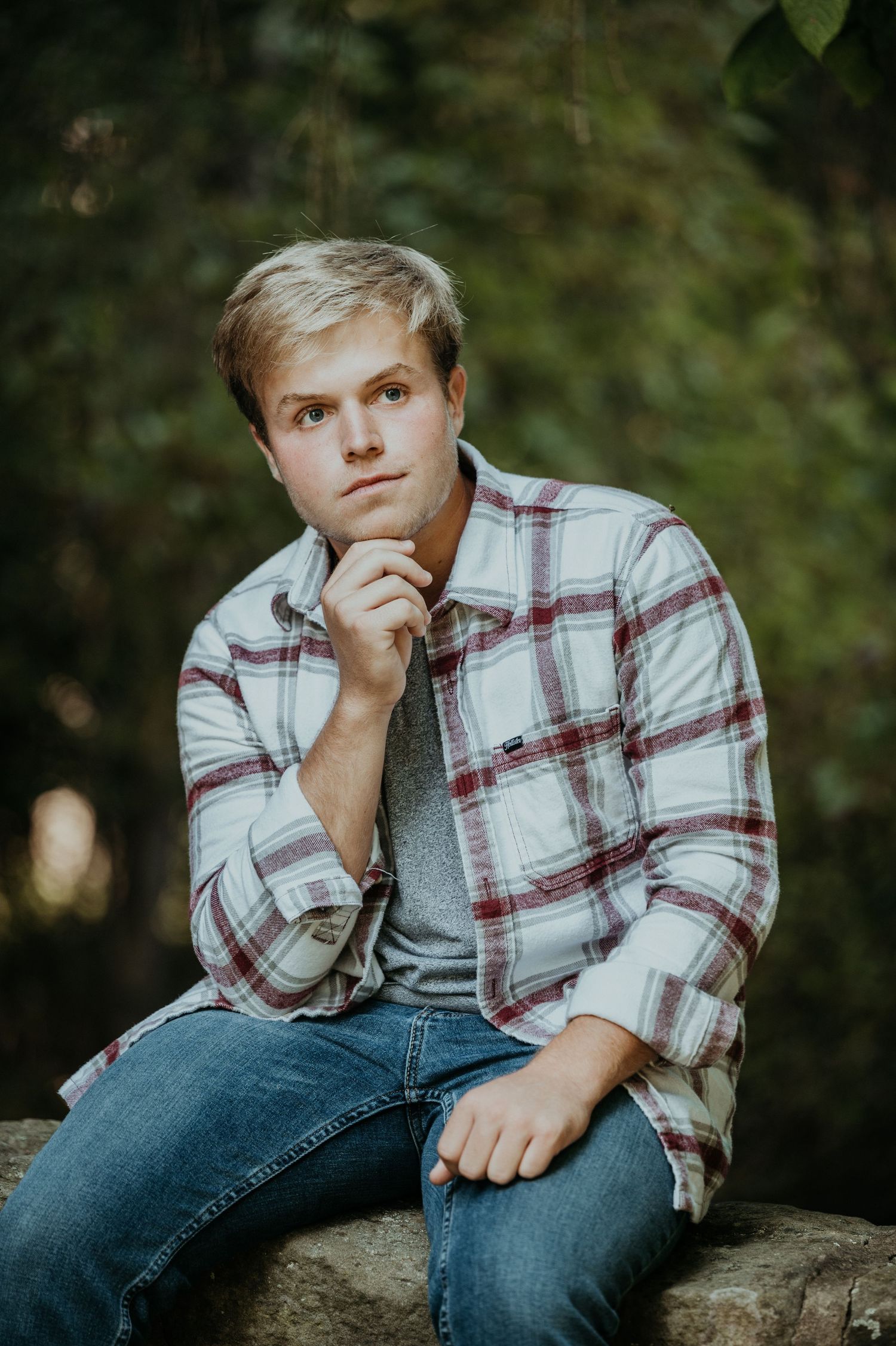 Young person in plaid shirt and jeans poses thoughtfully outdoors on a natural stone surface with greenery in background.