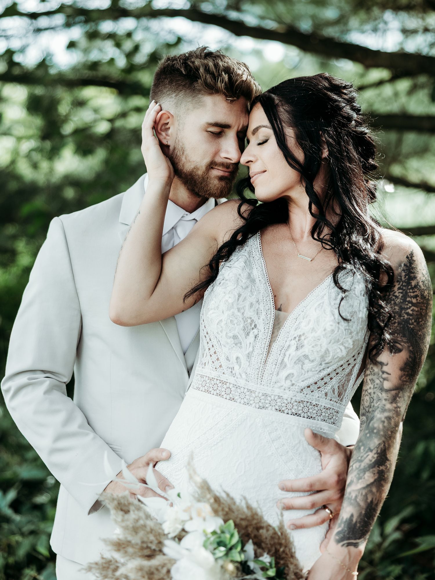 Intimate wedding couple embraces outdoors in elegant lace dress and light suit against green forest backdrop.