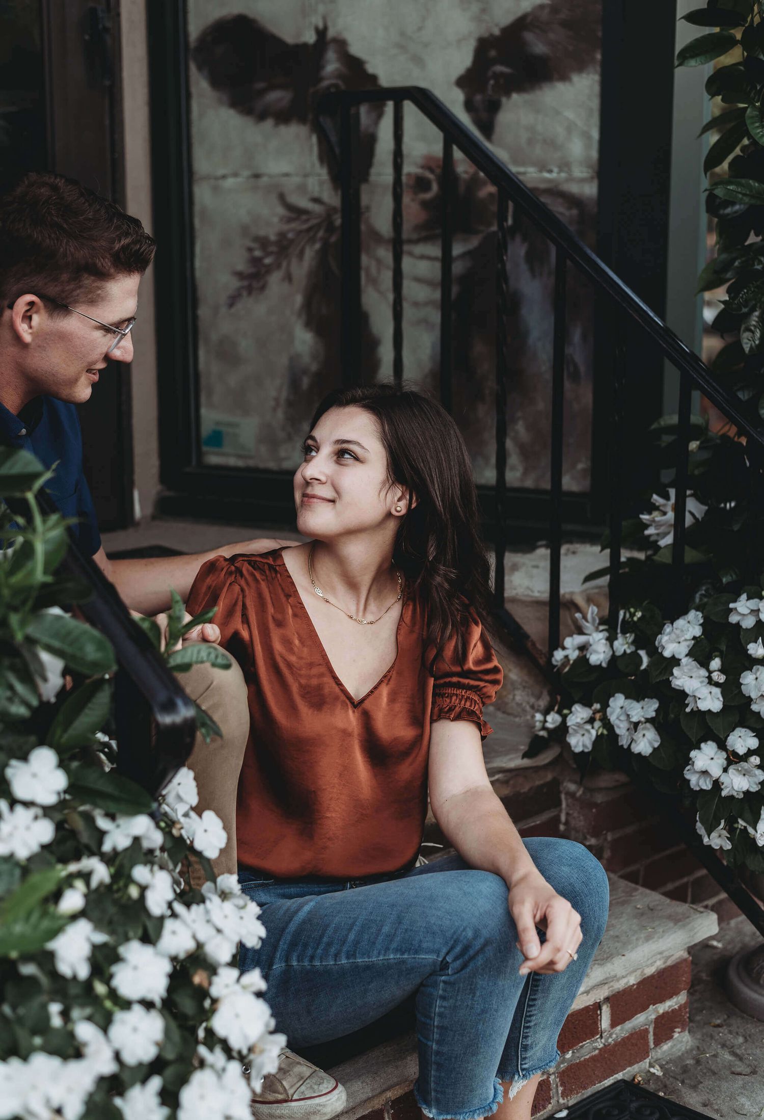 Woman in rust-colored blouse and jeans poses casually on brick steps with white flowers in garden setting.