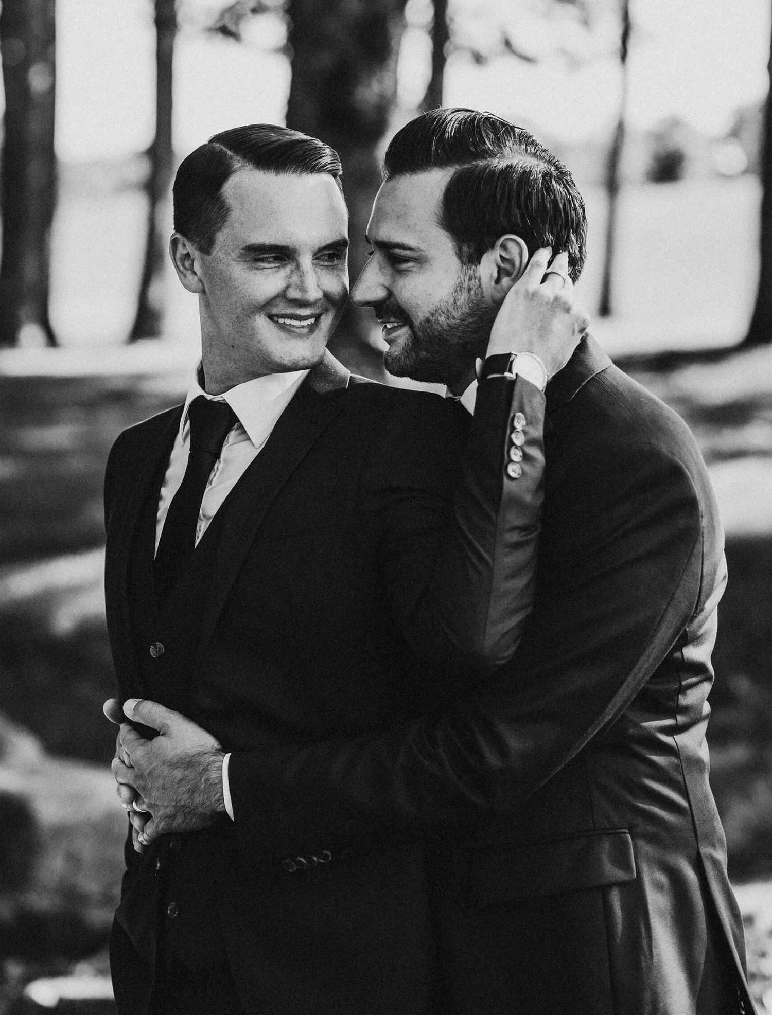A black and white photo of two people in suits sharing an intimate moment together outdoors.