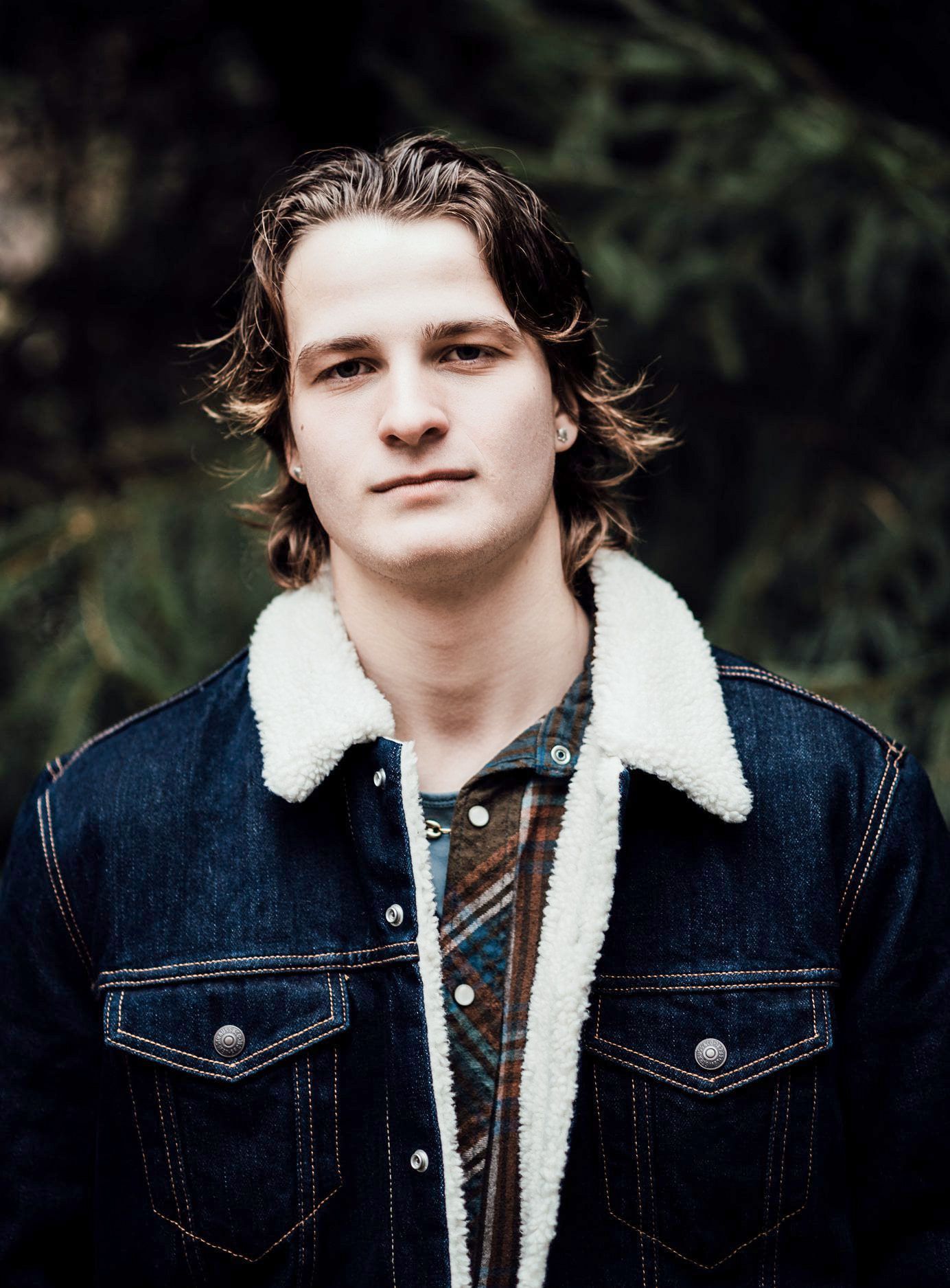 Person wearing a denim jacket with sherpa collar poses outdoors against a dark green background.