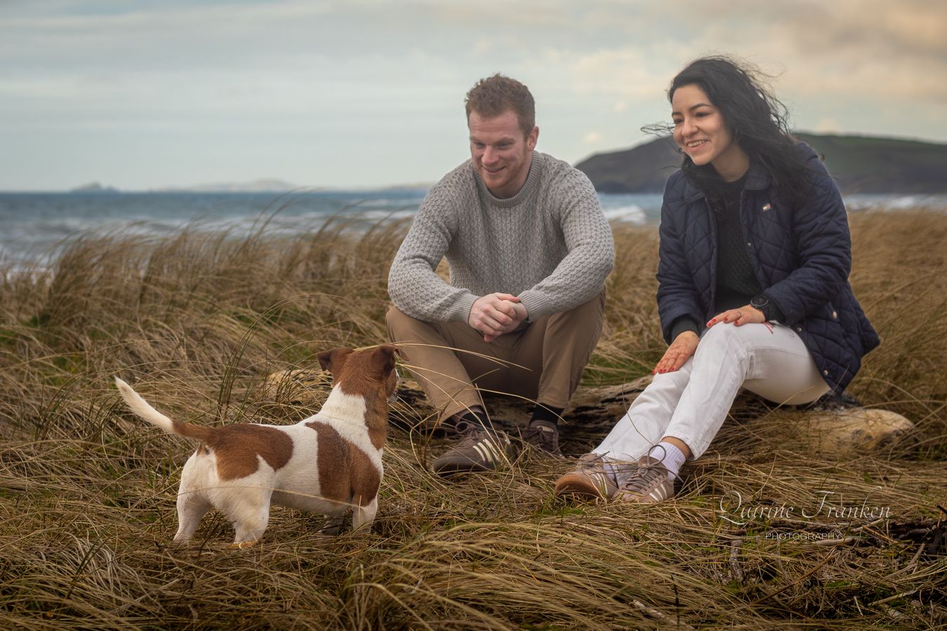 Couple sitting in the dunes looking a their dog during a photoshoot