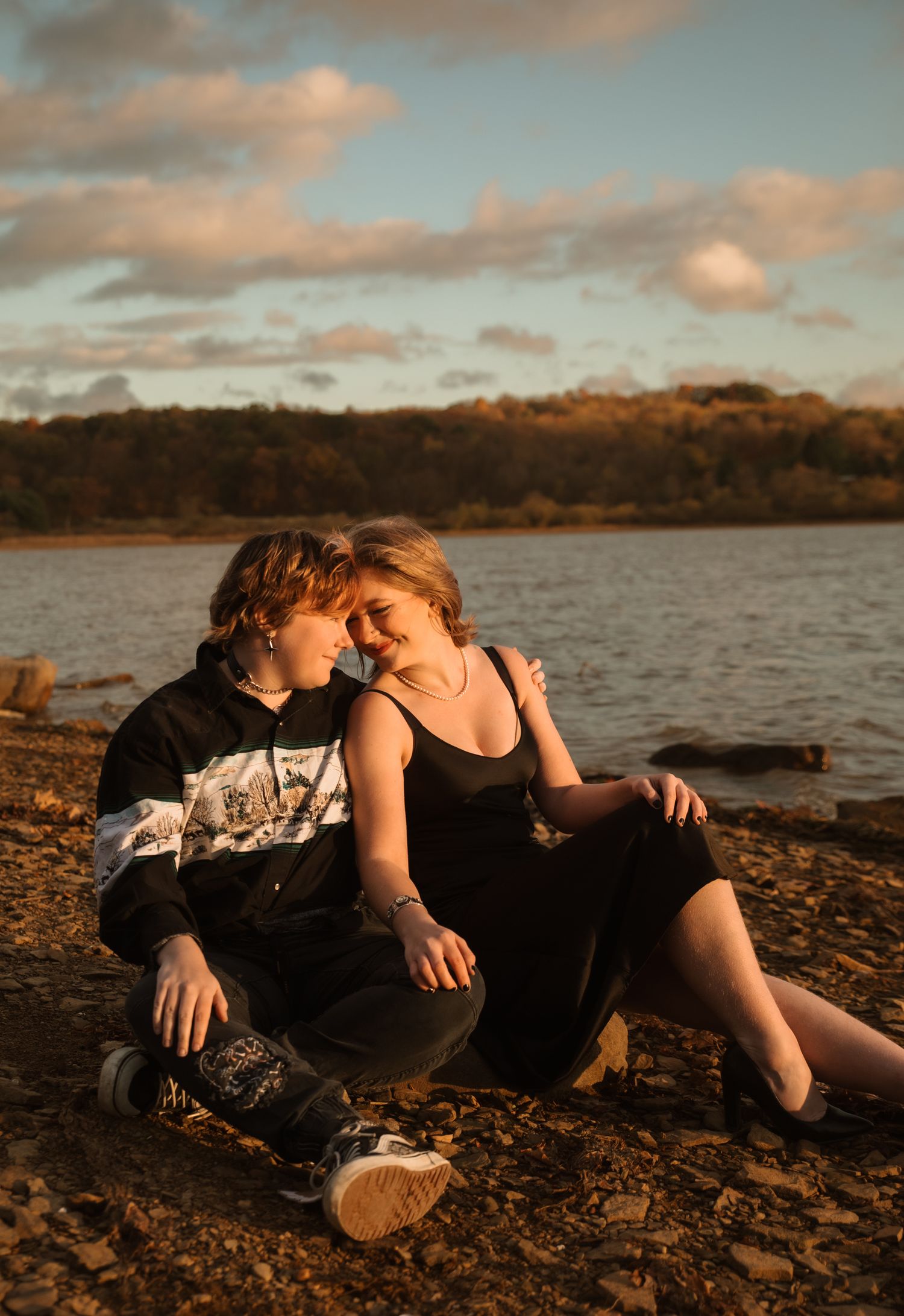Couple relaxing together on rocky shoreline at sunset with warm golden light reflecting off the water.