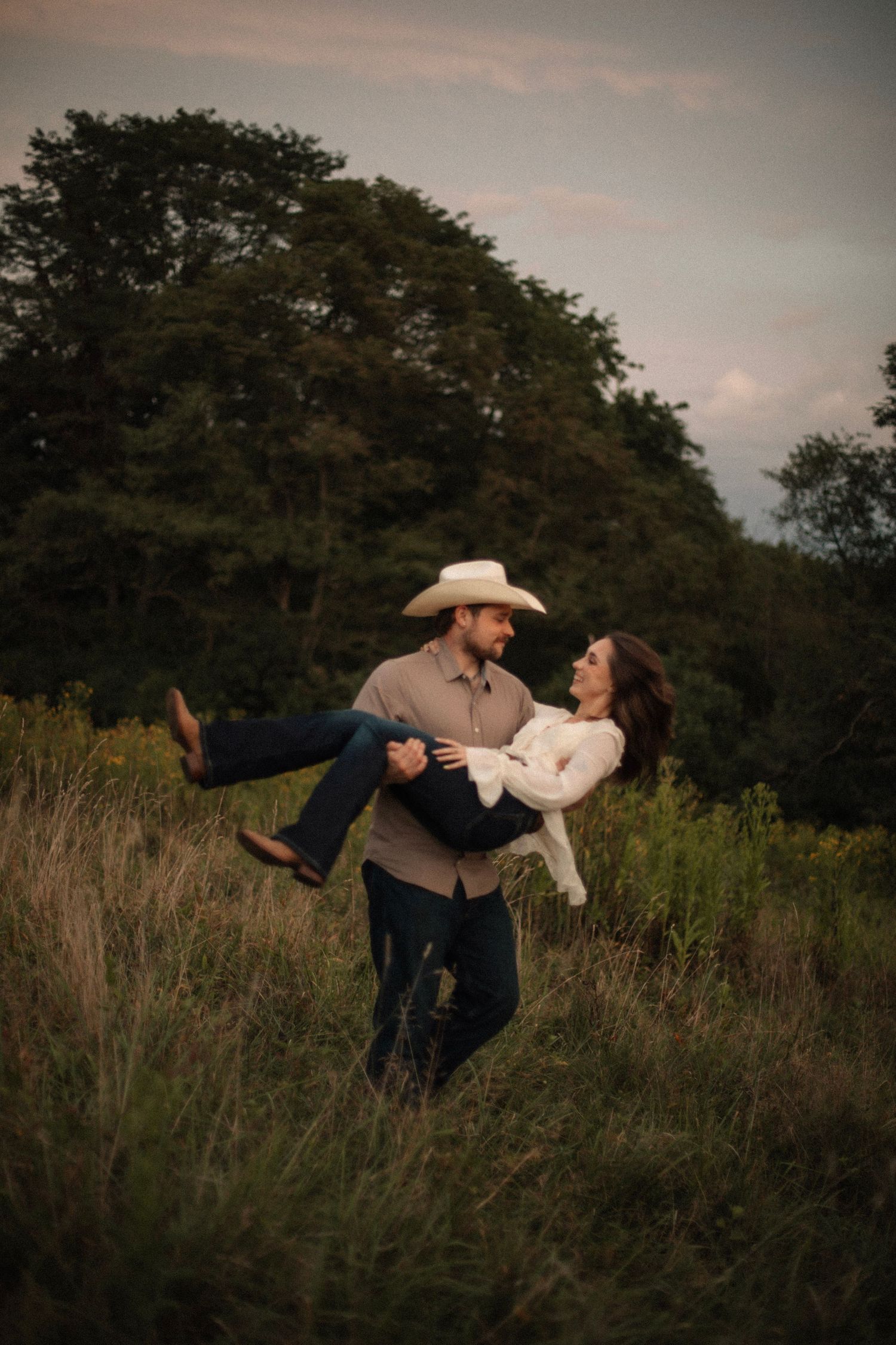 A man in a cowboy hat lifts a woman in a white blouse in a lush green field at dusk.