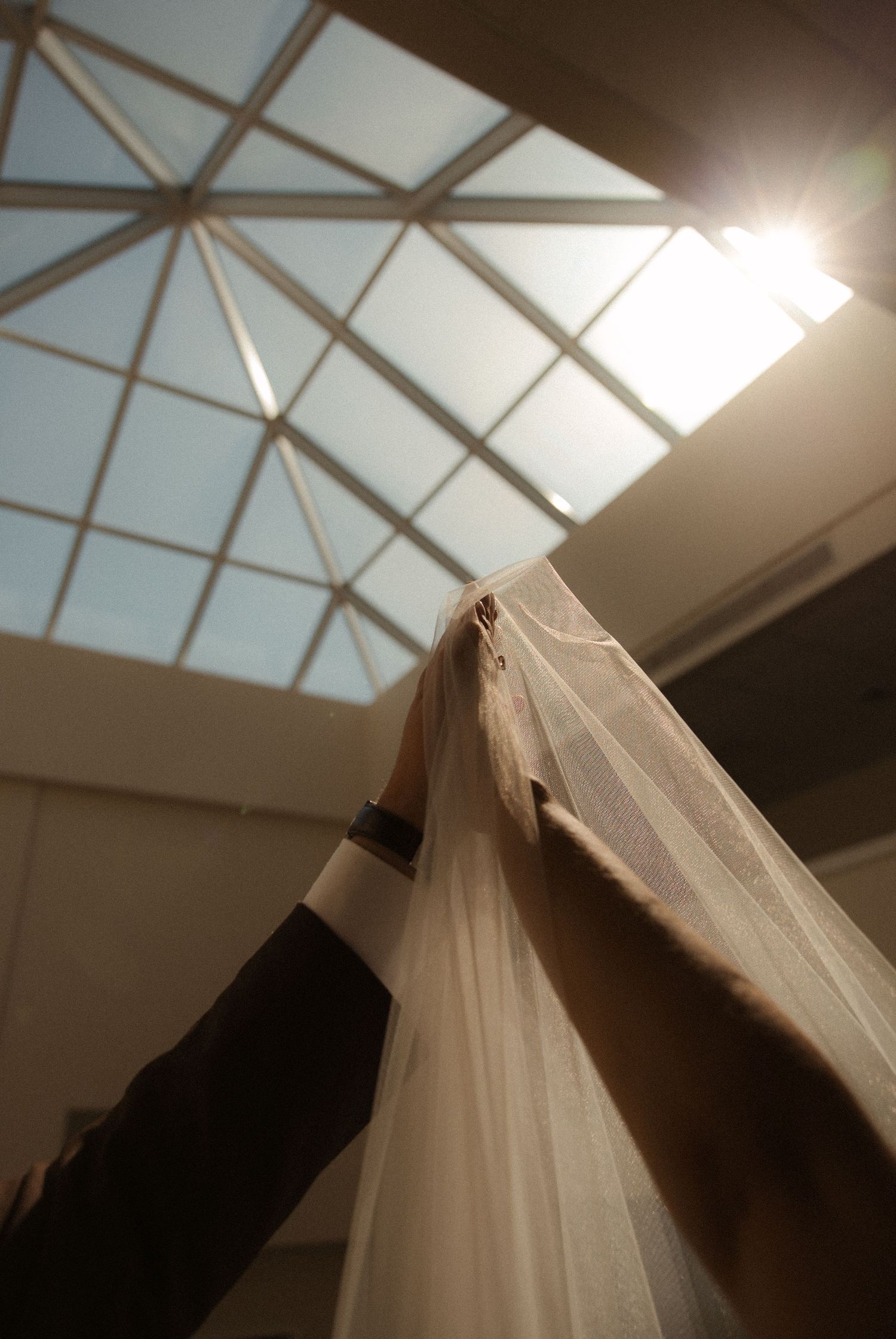 Hands holding a sheer white wedding veil beneath a sunlit geometric skylight ceiling.