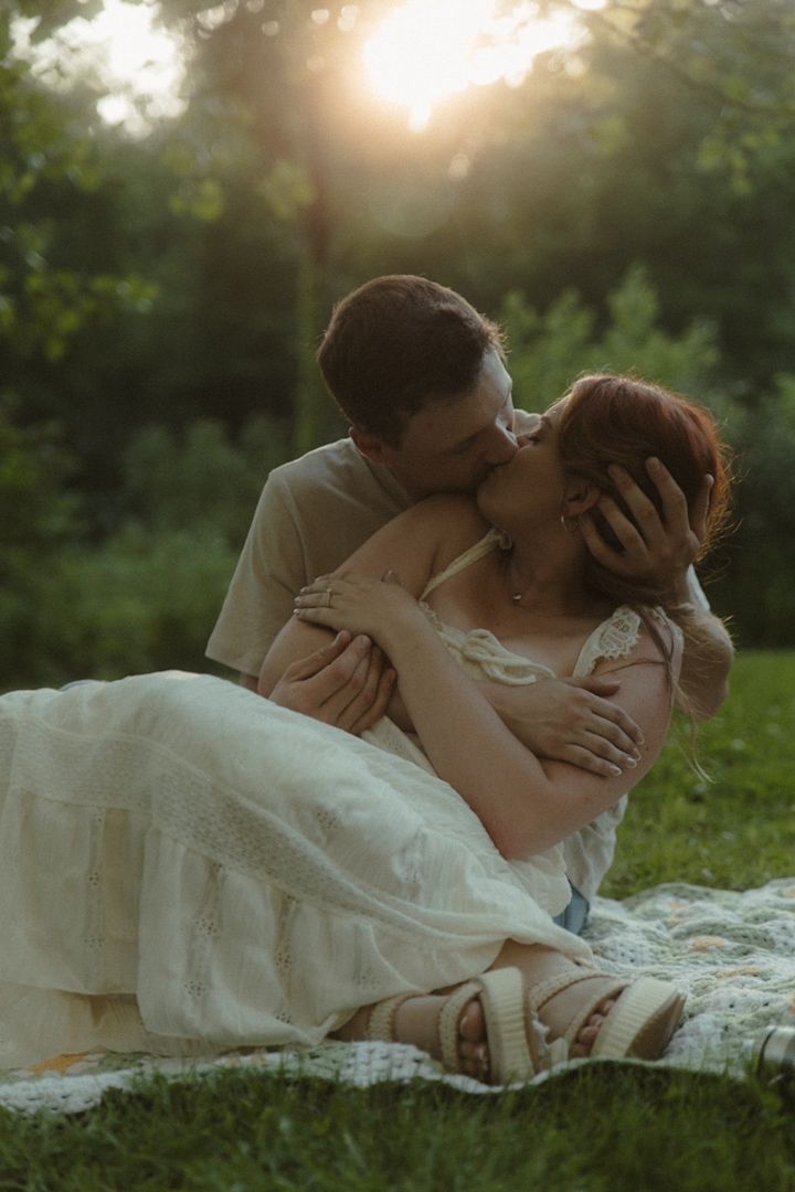 A couple shares a romantic kiss on a blanket in a sunlit garden, the bride wearing a white lace dress at golden hour.
