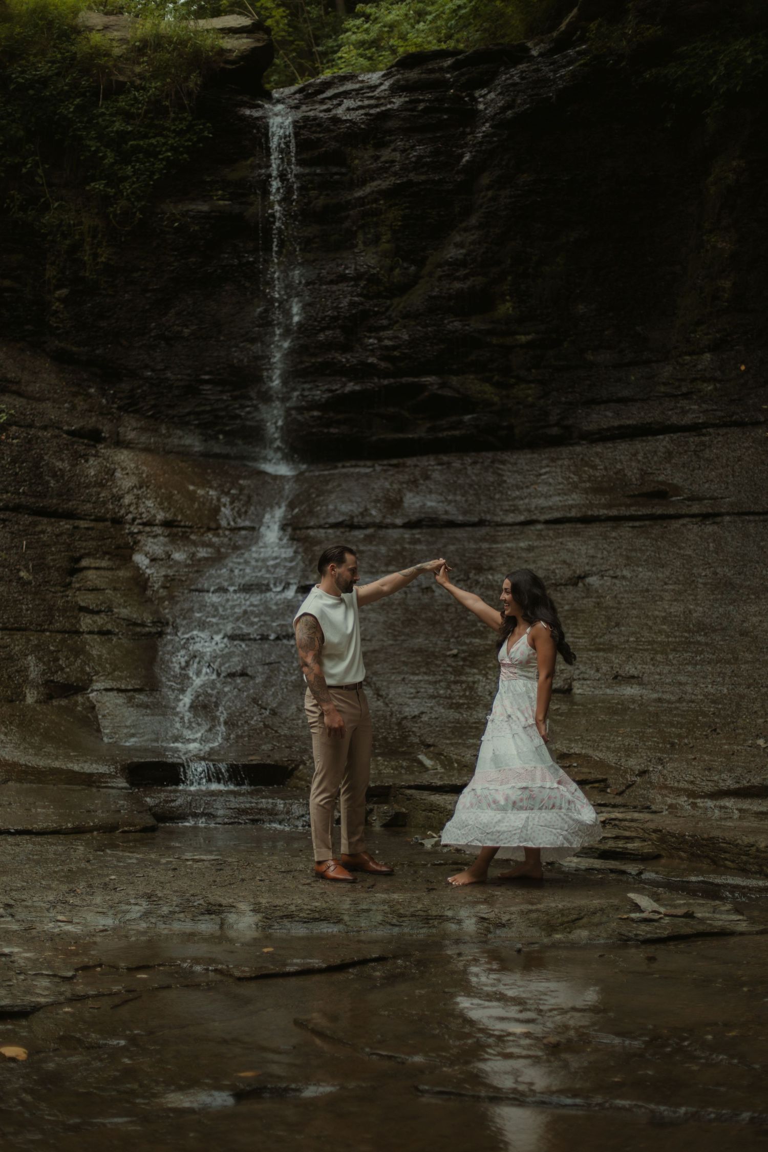 Couple dancing in front of a waterfall on rocky terrain, woman in white dress, man in tan pants, lush greenery above.