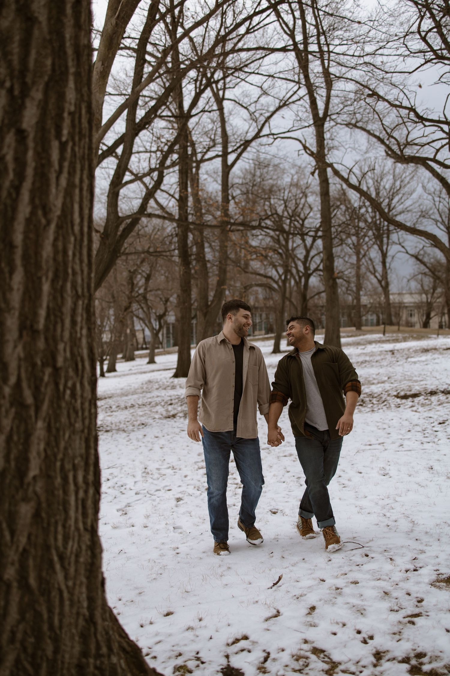 Two men holding hands and walking together through a snow-covered park with bare trees on a cloudy winter day.
