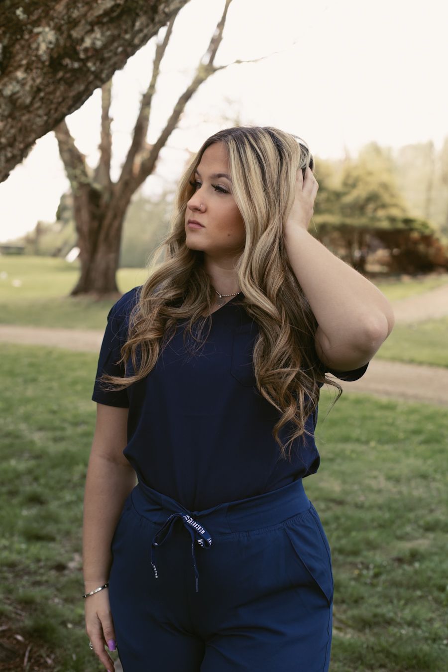 Woman in navy blue scrubs poses outdoors near trees, touching her highlighted wavy hair in soft natural light.