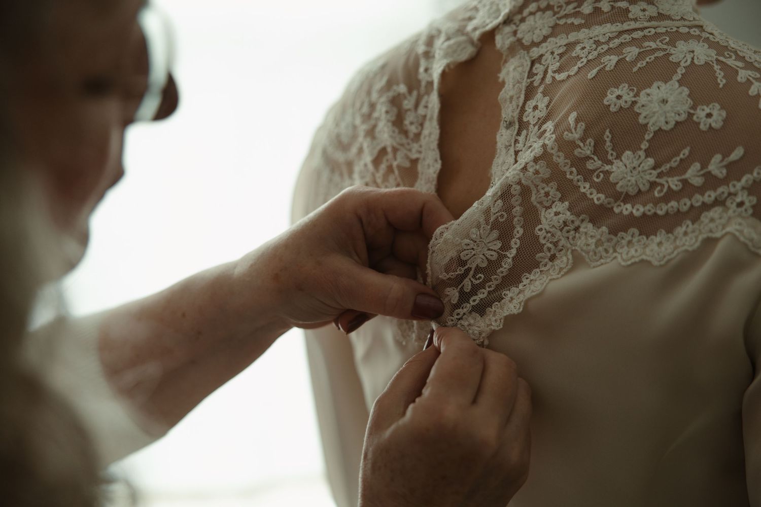 A bridesmaid carefully fastening delicate lace buttons on the back of a bridal wedding gown.