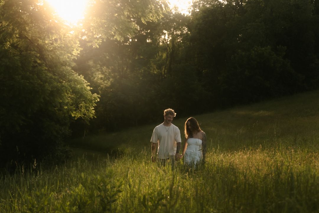 Couple holding hands walking through a sunlit green meadow surrounded by lush trees during golden hour sunset.