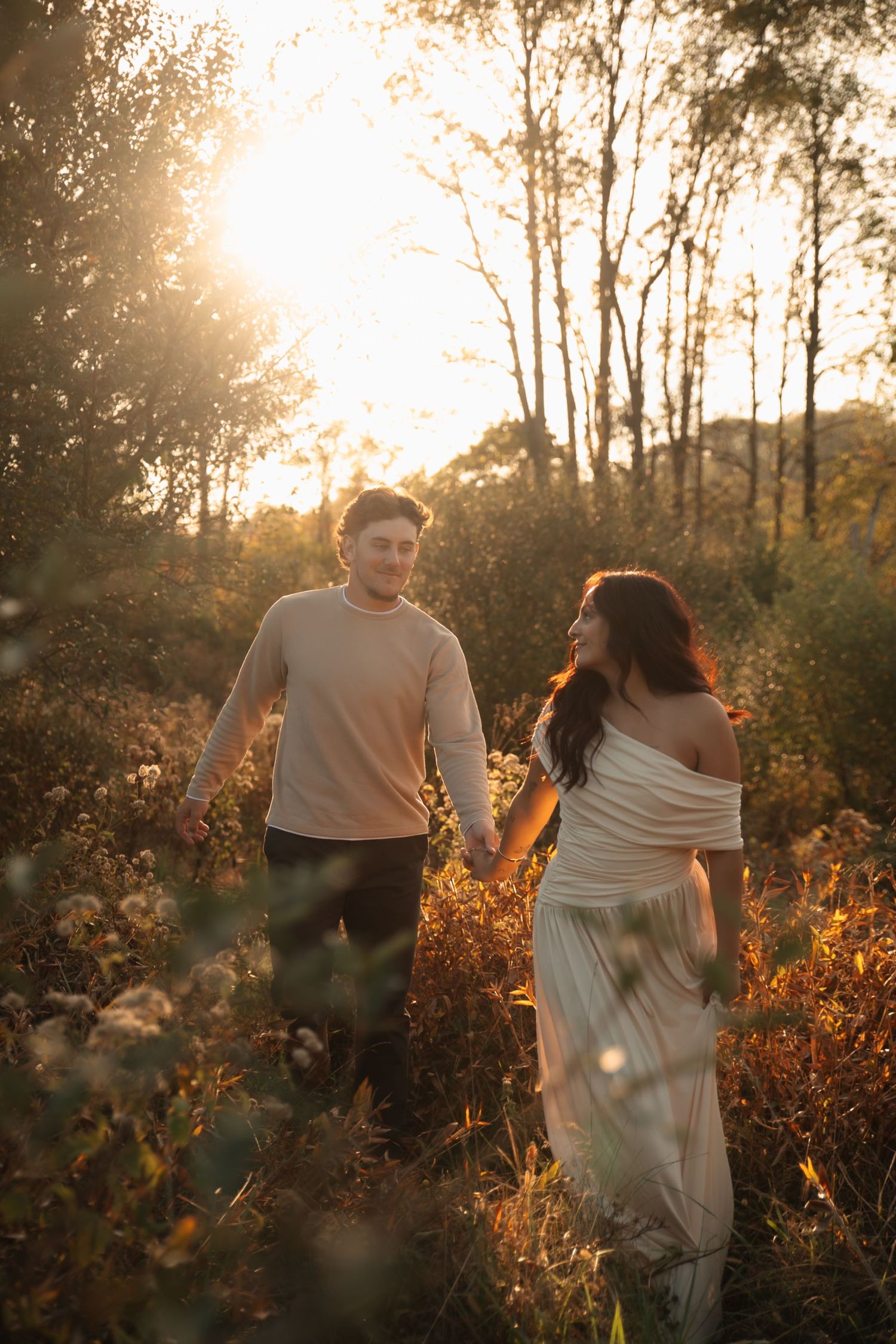 Couple holding hands walking through golden wildflower field at sunset, woman in white off-shoulder dress, man in beige sweater.