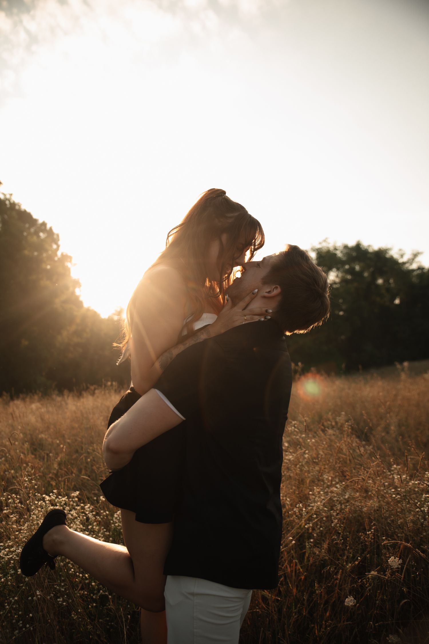 A couple shares a romantic kiss at sunset in a golden field, with warm backlit sunlight glowing behind them.