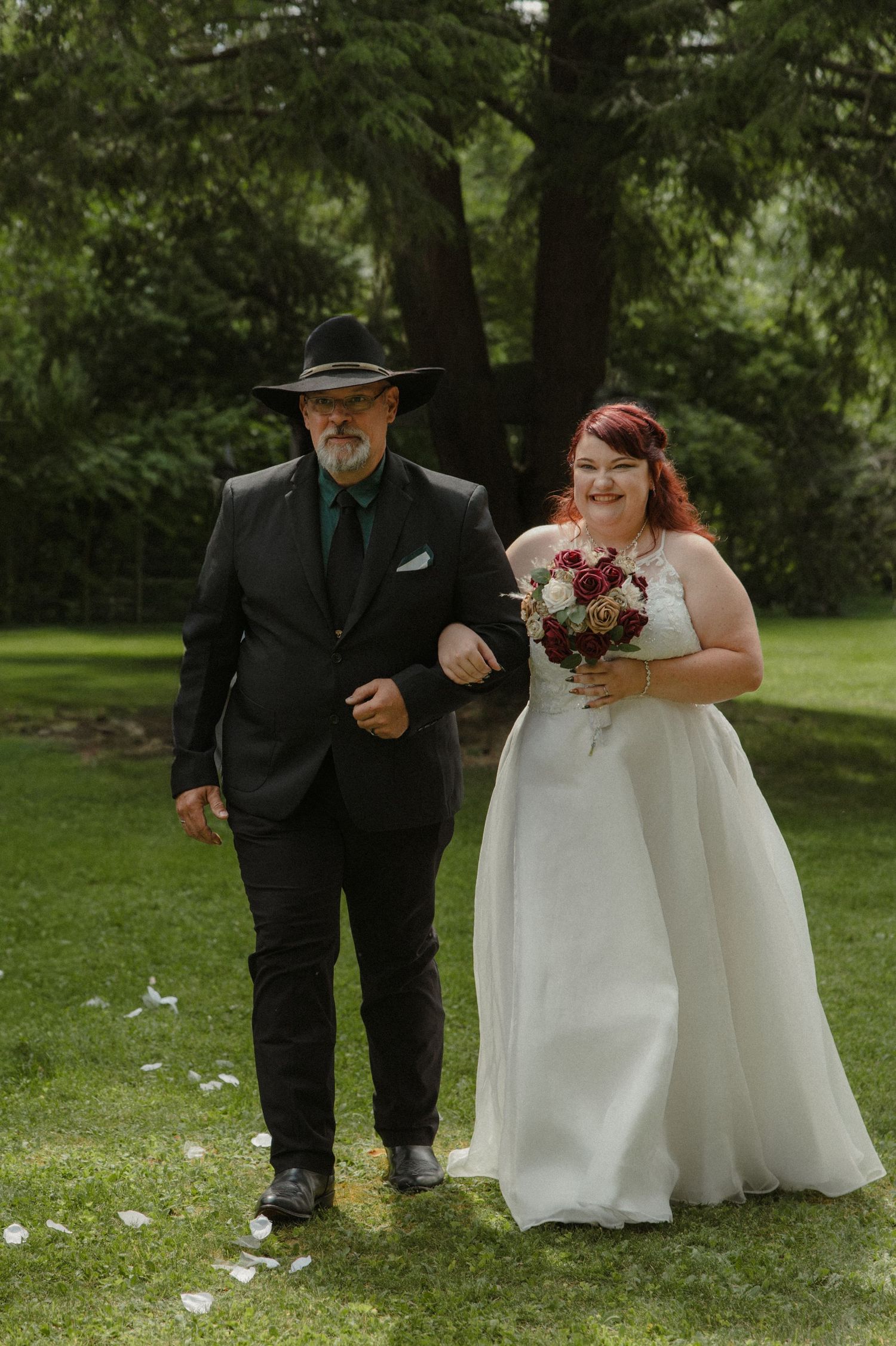 Bride in white gown holding bouquet walks down outdoor aisle with man in black suit and wide-brim hat.