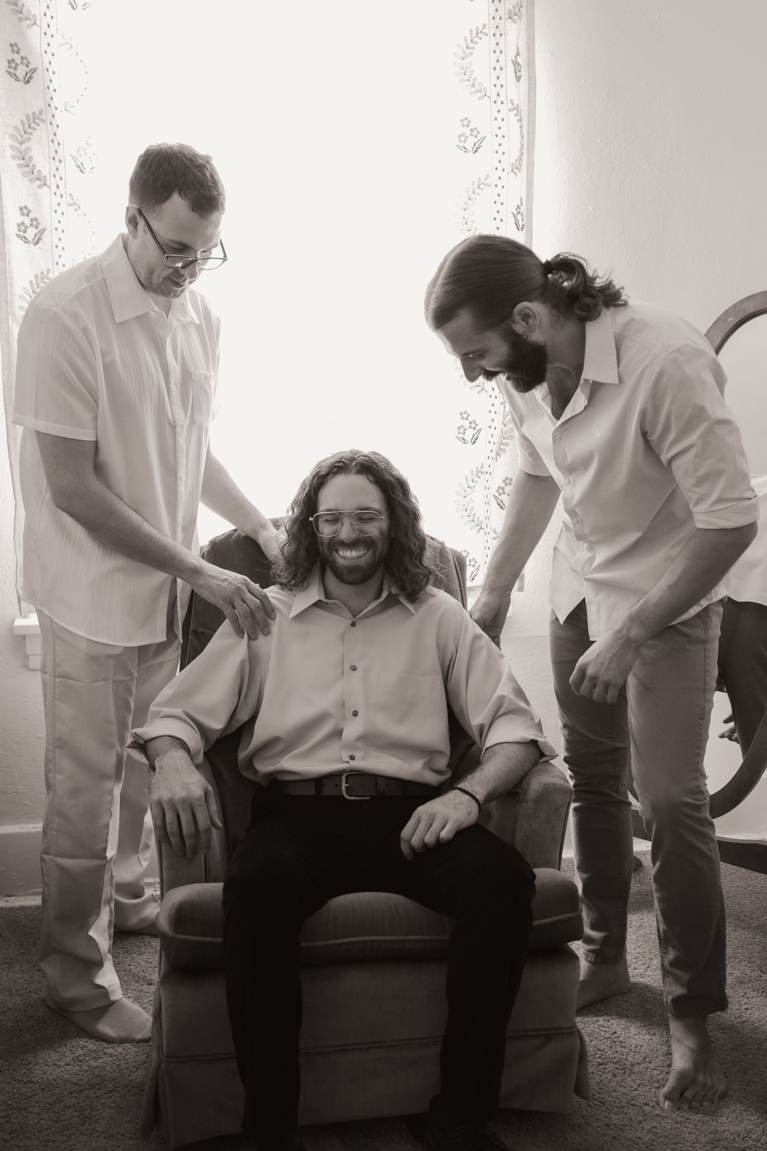 Three men smiling in black and white photo, two standing beside a seated man with curly hair in a bright room.