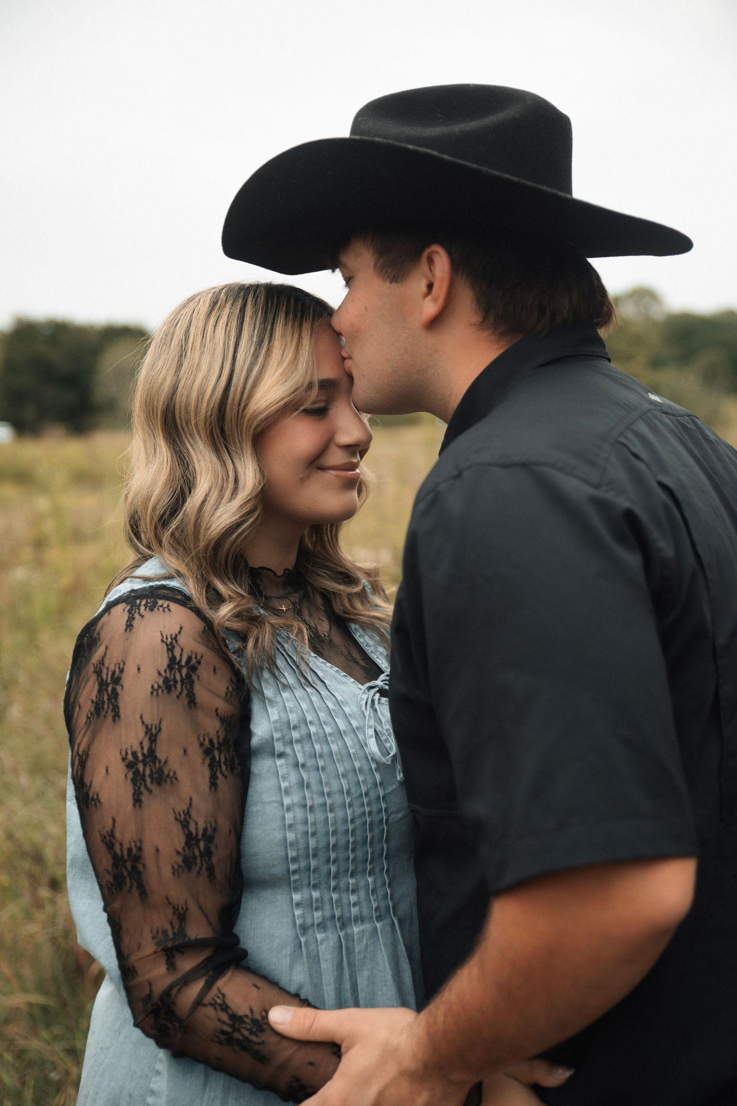 A romantic couple embraces in a field, with her wearing a blue dress with black lace sleeves and him in a cowboy hat.