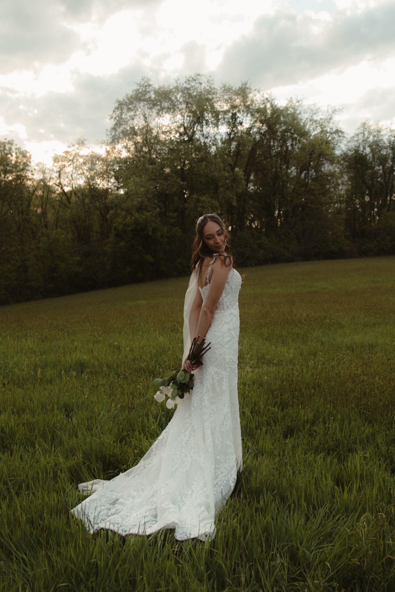 Bride in white lace gown holding bouquet stands in green field at sunset with trees in background.