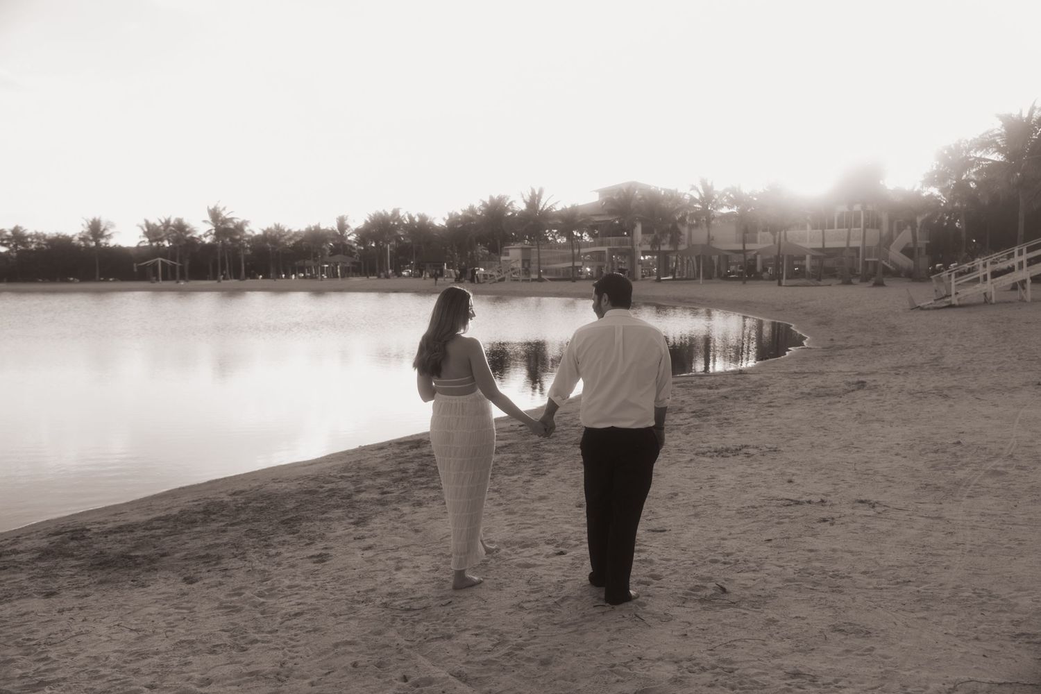A couple walks hand in hand along a peaceful lakeside path on a serene afternoon in black and white.