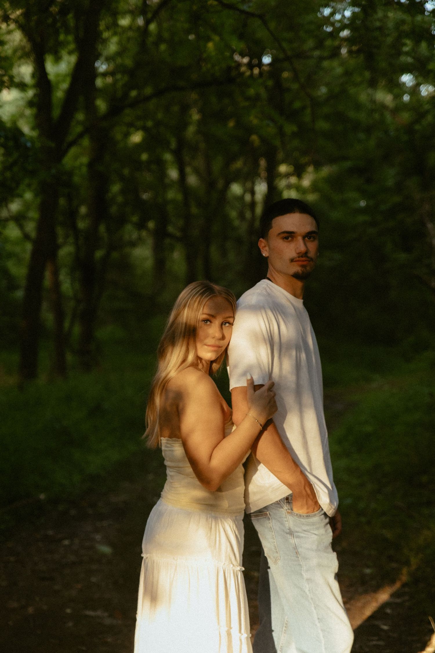 A couple poses in a sunlit forest, woman in white dress embracing man in white shirt and jeans on a shaded path.