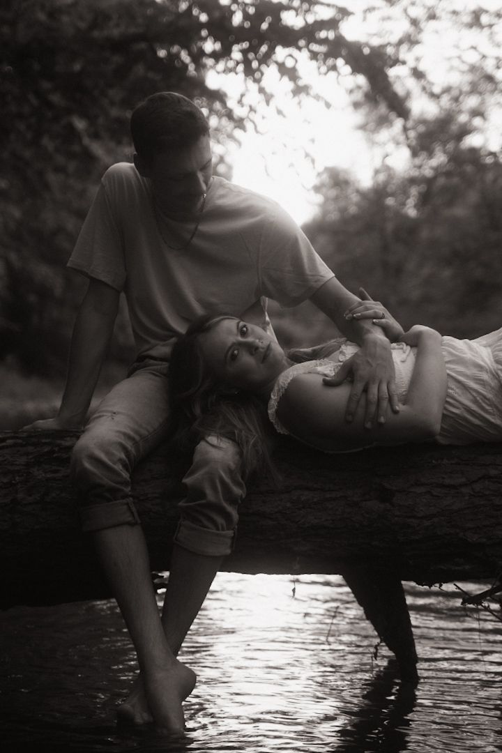 Black and white photo of a couple sitting on a log over water, woman reclining against man in romantic forest setting.