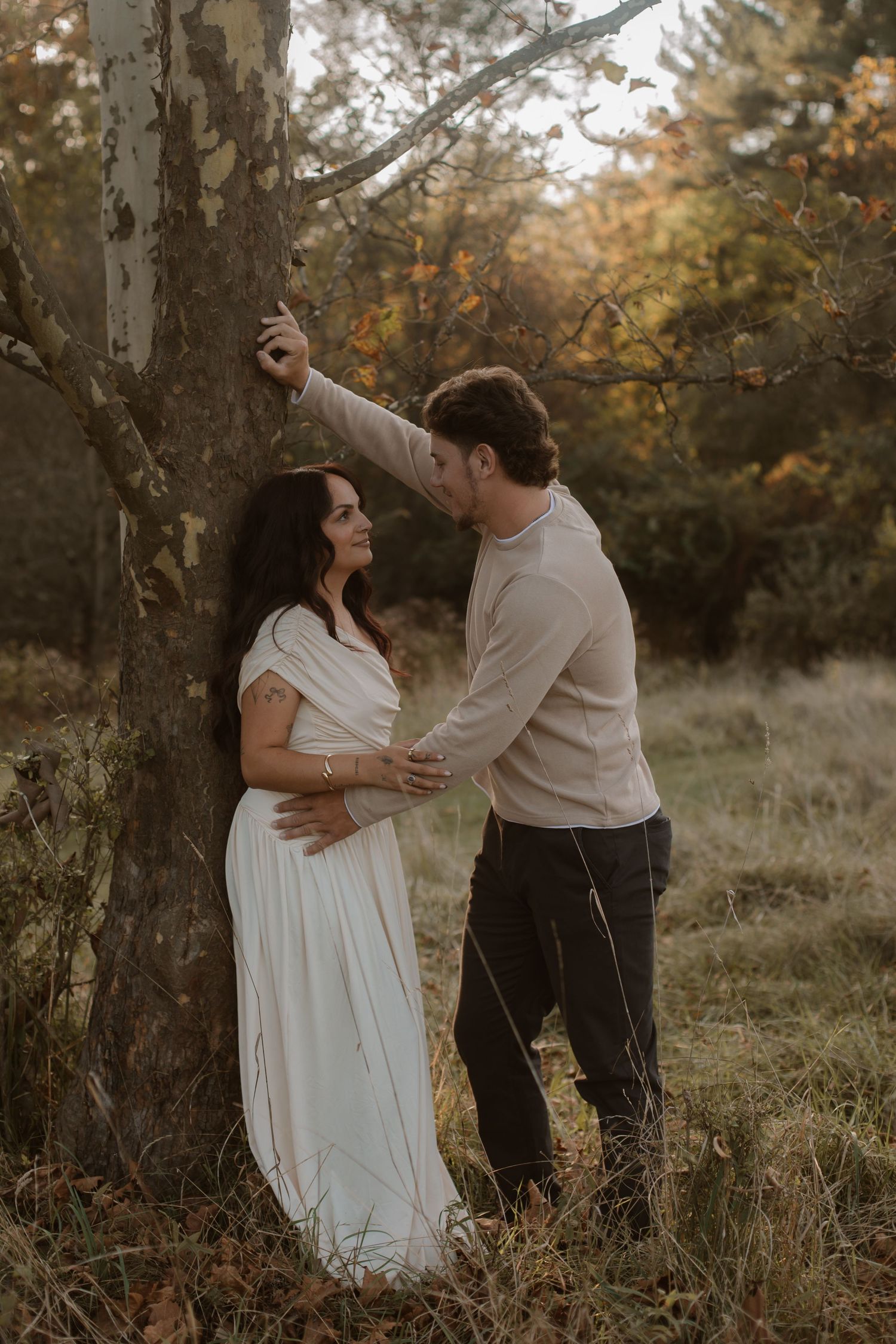 Couple sharing a tender moment by a tree in autumn, woman in white maternity gown, man in beige sweater.
