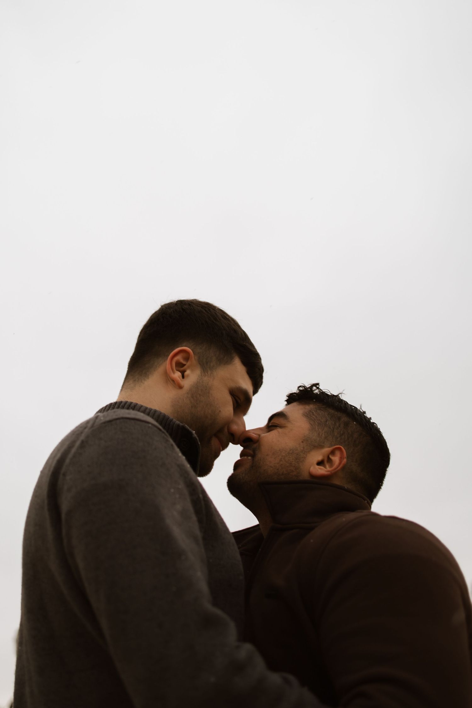 Two men sharing a tender kiss outdoors against a grey sky, wearing casual sweaters in an intimate moment.