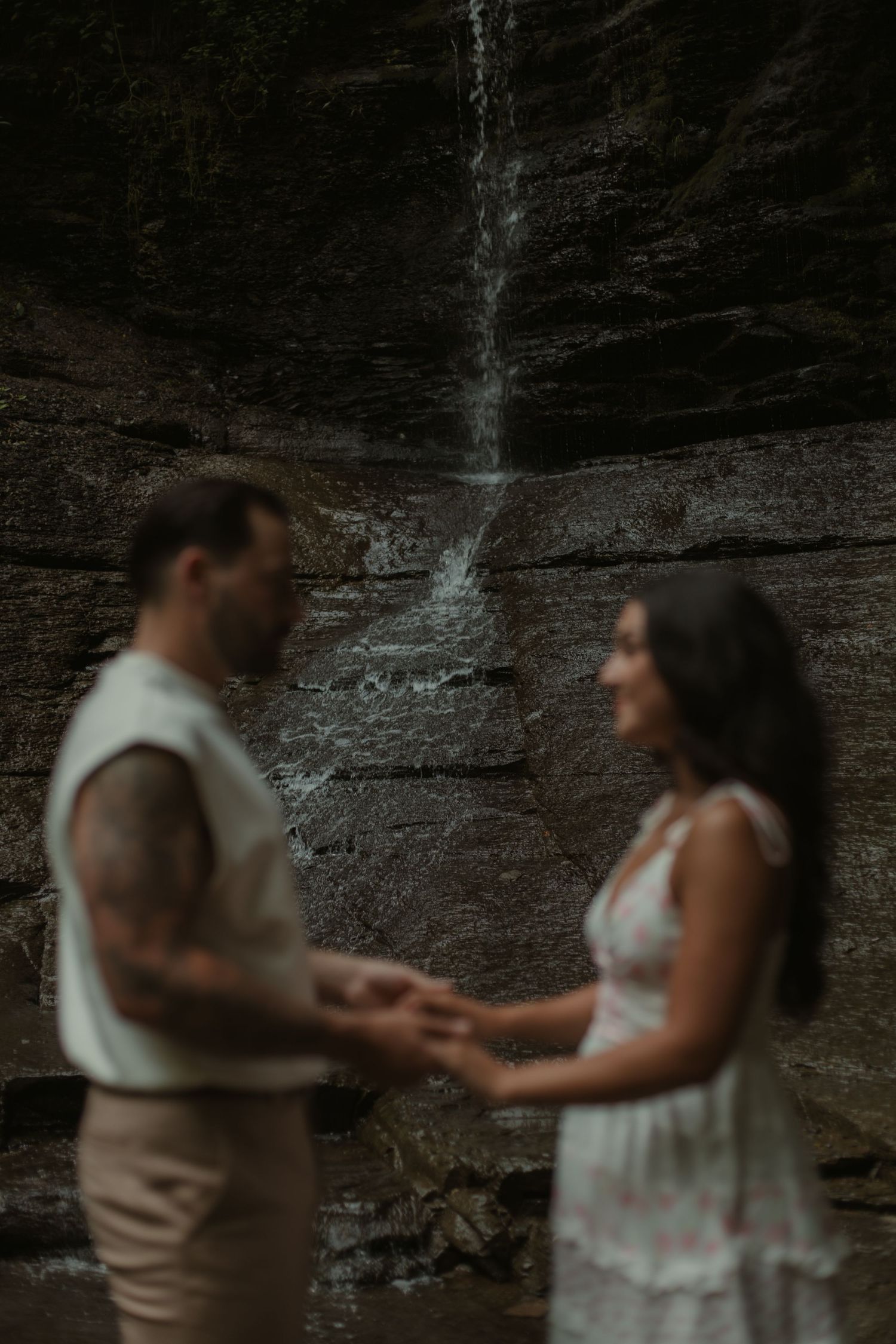 Couple holds hands in front of a waterfall cascading down rocky cliffs in a romantic outdoor portrait.