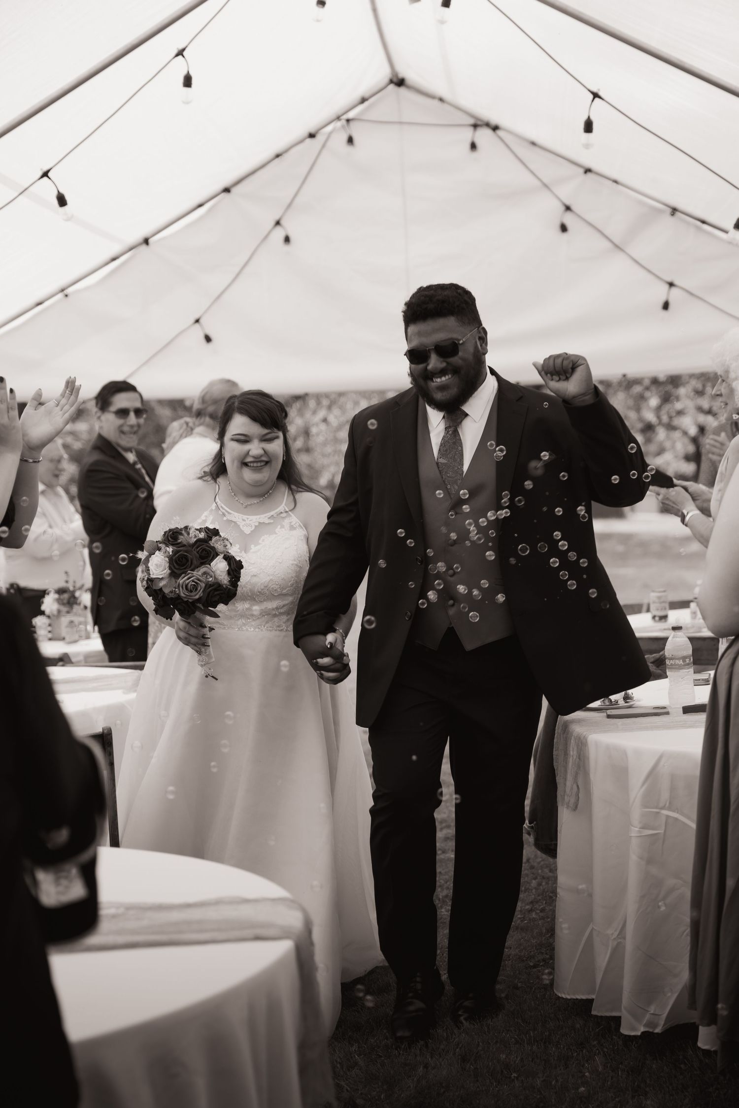 Black and white photo of a joyful newlywed couple walking under a tent at their outdoor wedding reception.