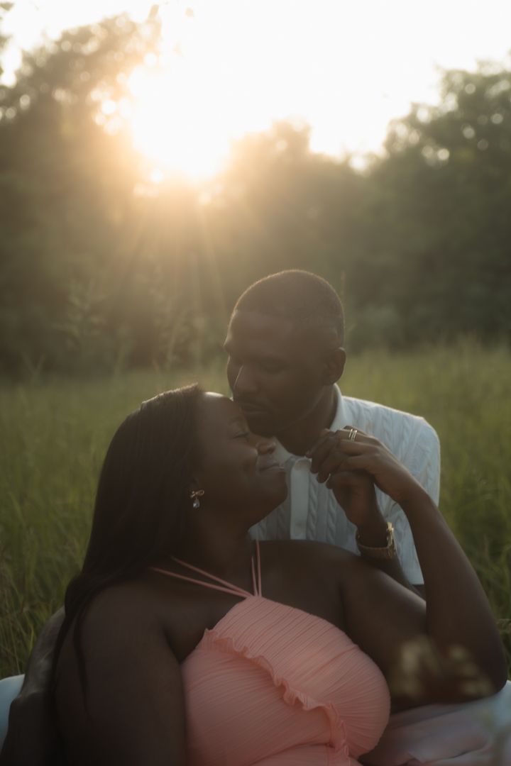 A couple shares a romantic moment at golden hour in a lush green field, with warm sunlight glowing behind them.