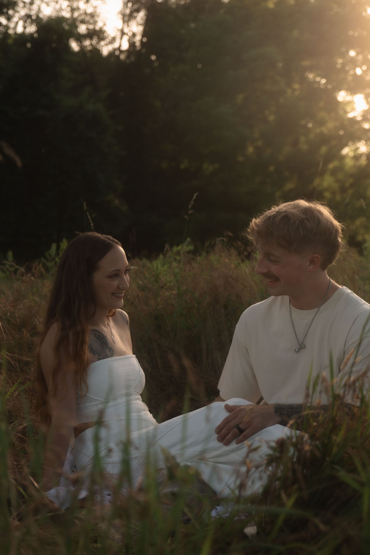 A couple sits together in a sunlit meadow at golden hour, smiling at each other surrounded by tall grass and trees.