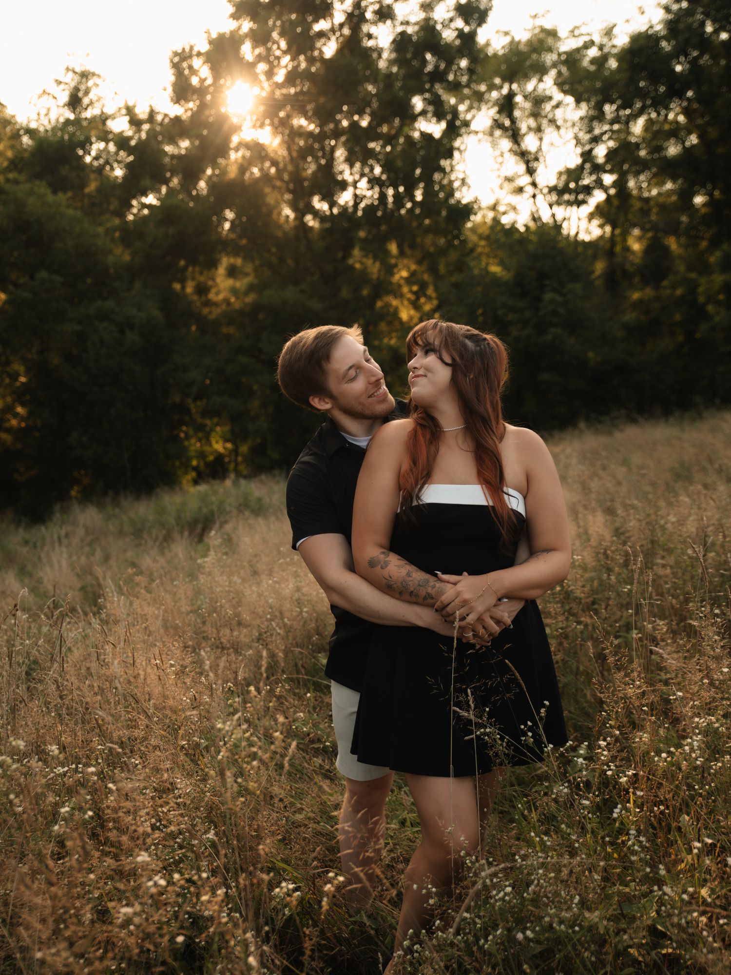 A couple embraces in a golden field at sunset with sunlight filtering through trees in the background.