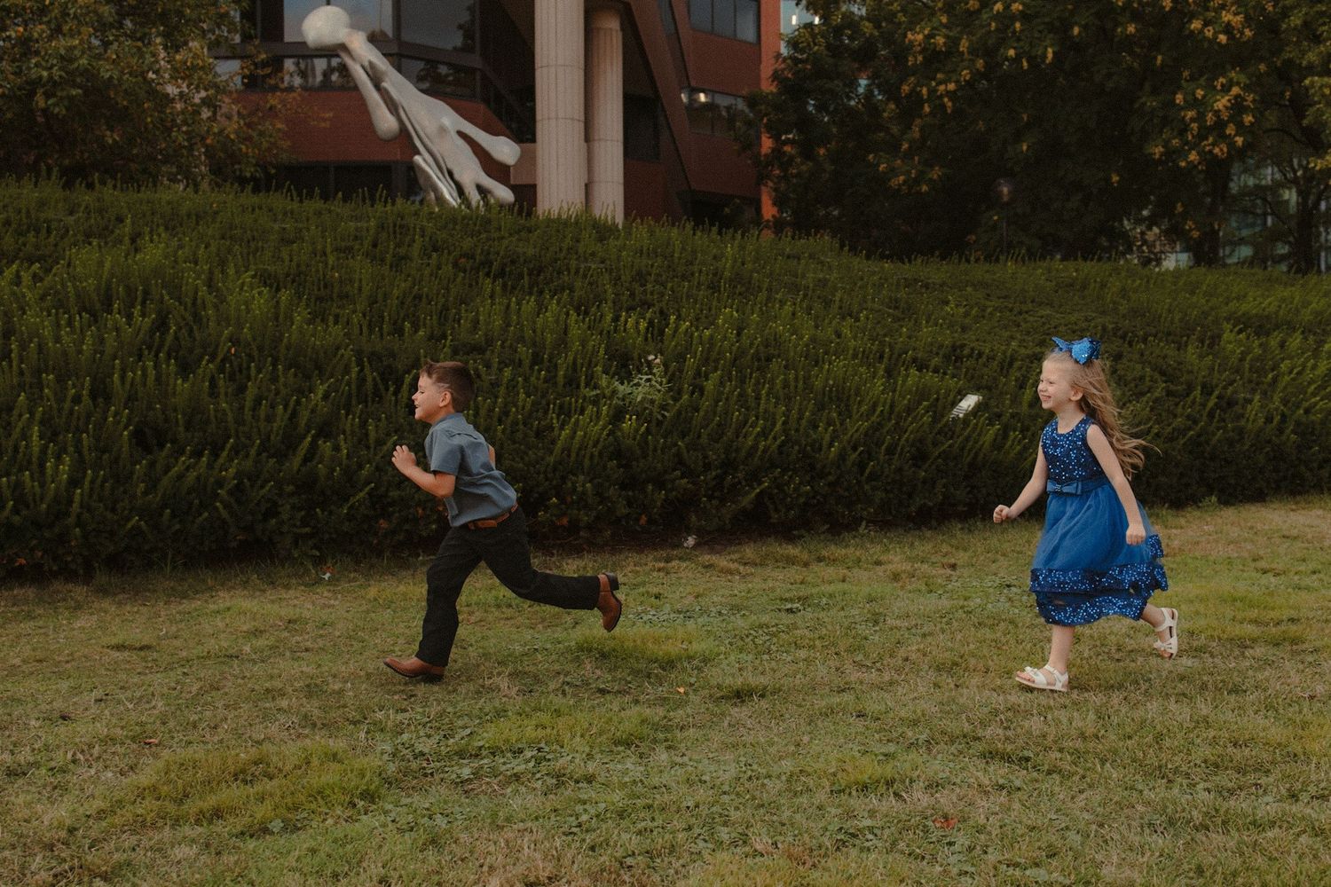 Two children in formal attire joyfully chasing a badminton shuttlecock on a green lawn near a building.