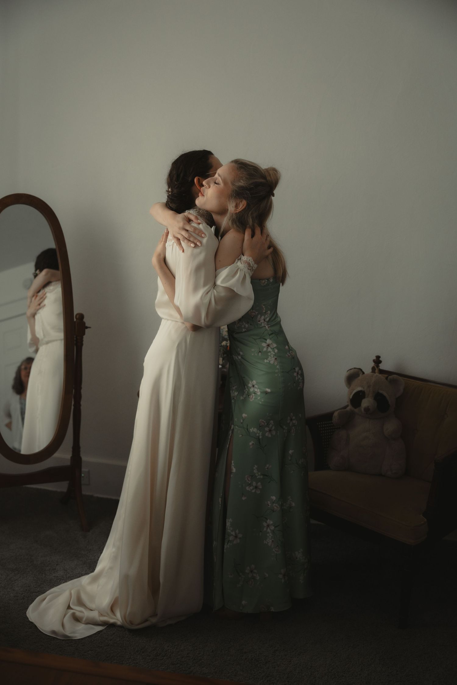 Bride in white gown embracing bridesmaid in green floral dress, sharing a tender moment near a mirror.