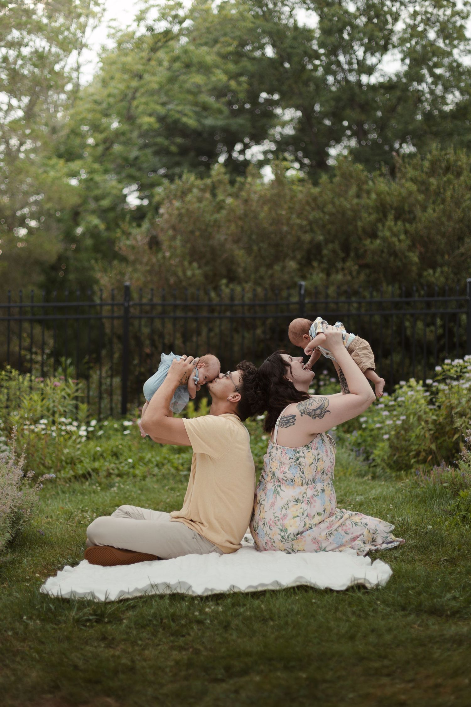 A couple sits back-to-back on a blanket in a garden, each holding a baby up in the air, smiling joyfully.