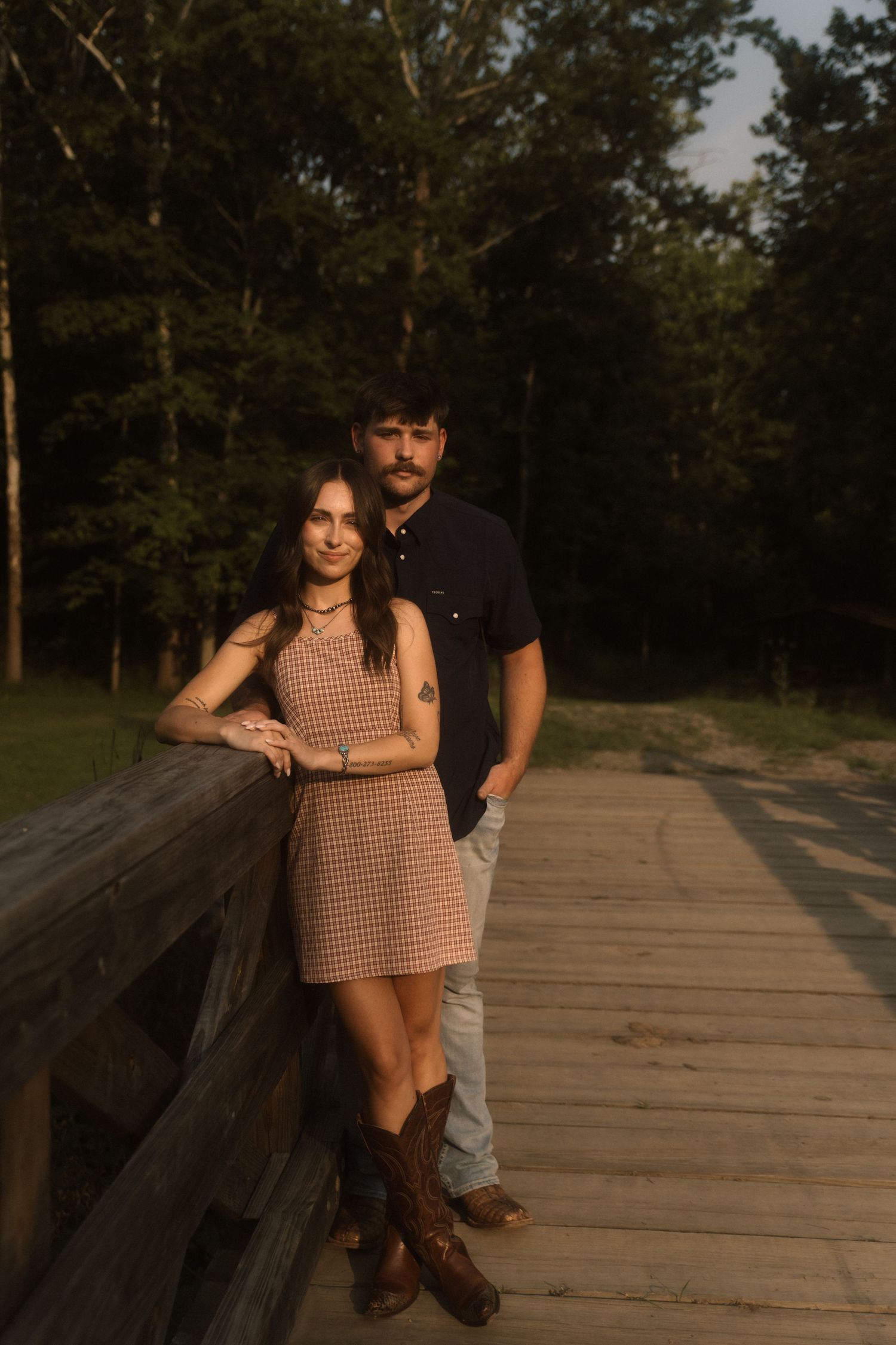 Couple posing on a wooden bridge at sunset, surrounded by lush green trees in a serene outdoor setting.