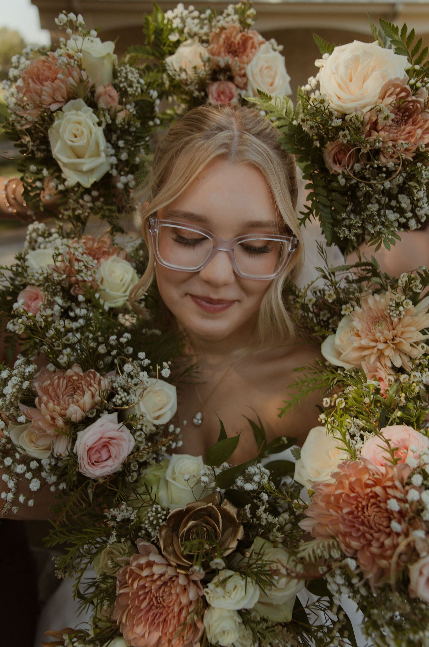 Bride surrounded by lush floral arrangement of blush roses, dahlias, and greenery, wearing clear-frame glasses.