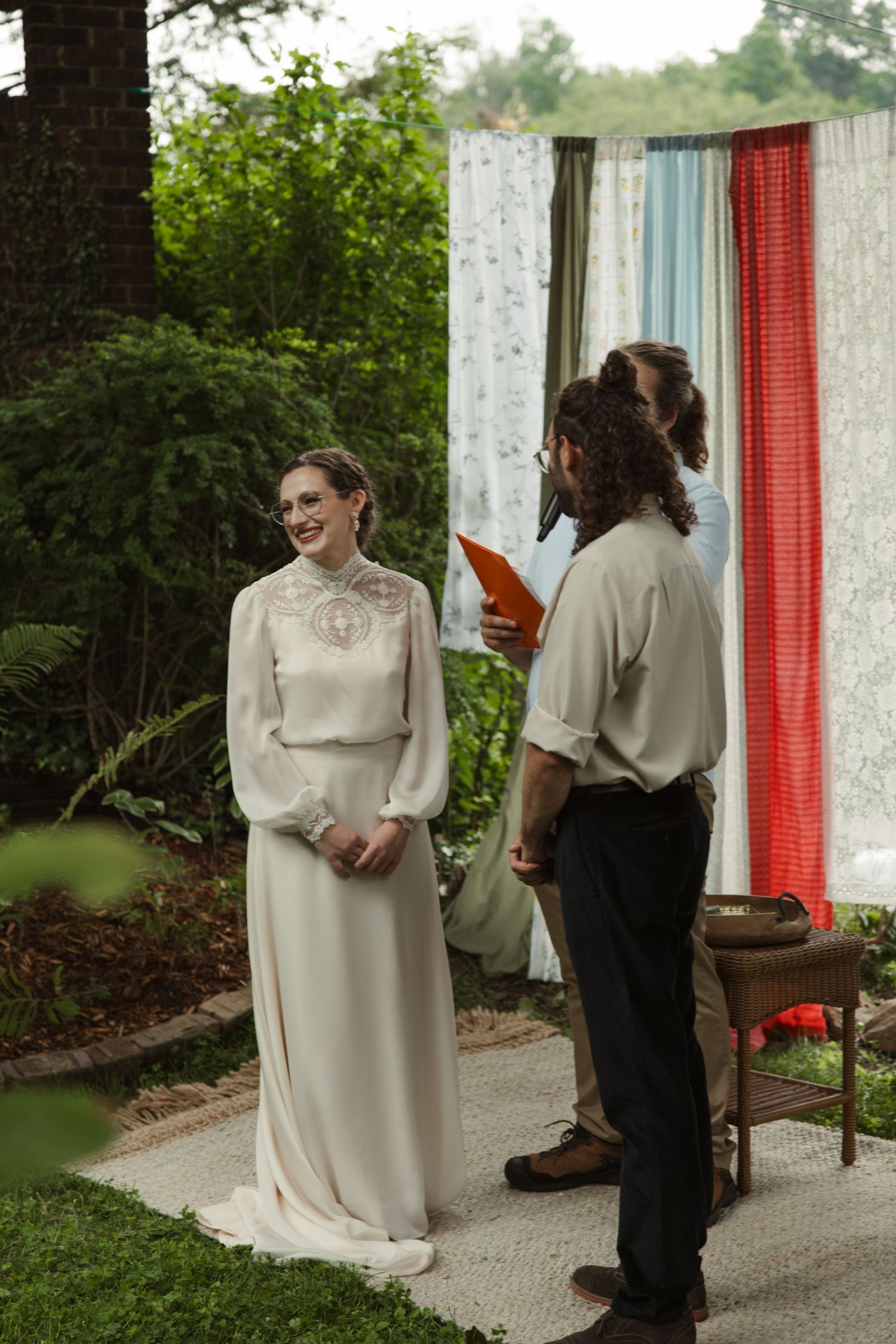Bride in white long-sleeve gown smiling during outdoor wedding ceremony with colorful fabric backdrop.