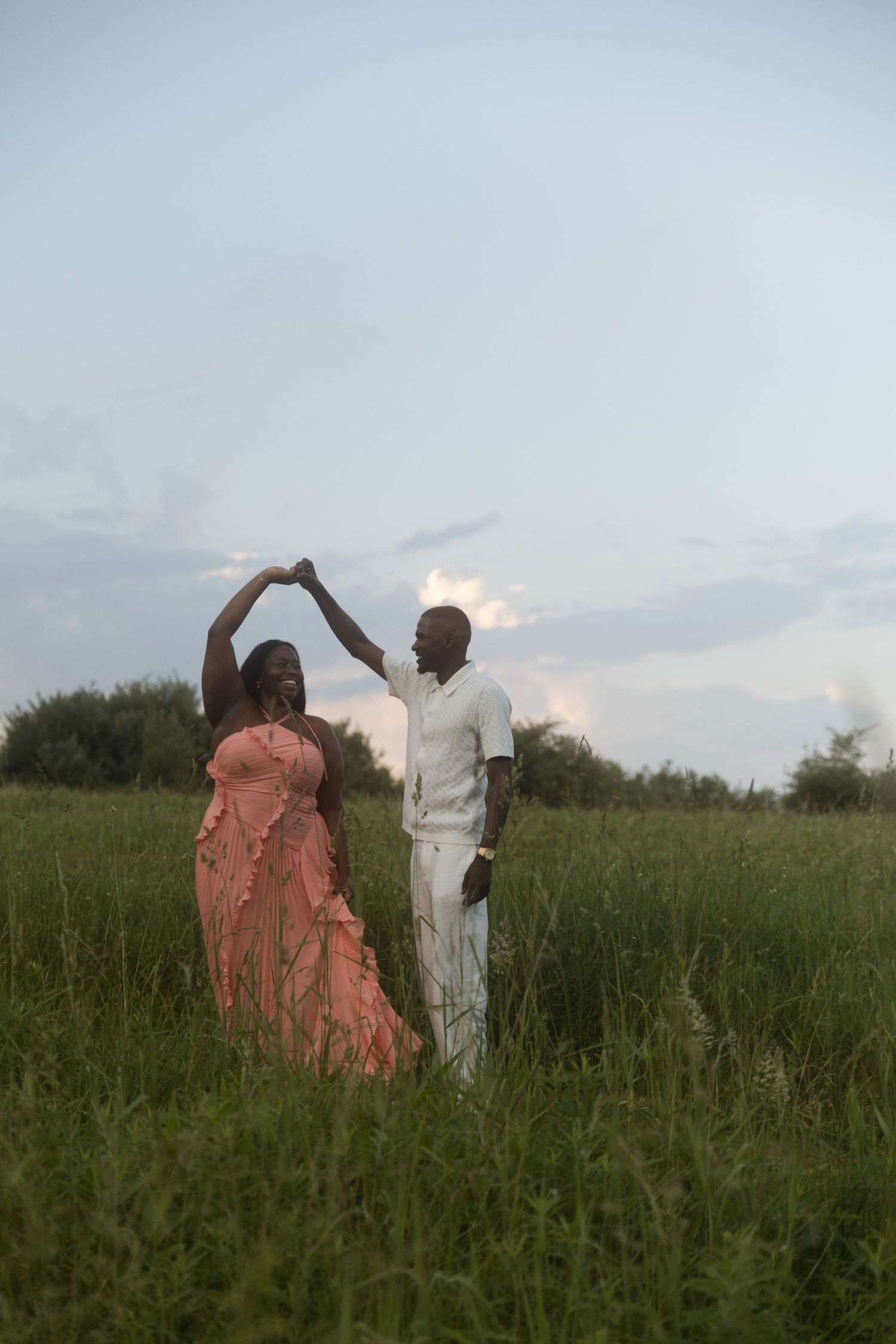 A couple dances romantically in a lush green field at dusk, woman in pink dress, man in white outfit.