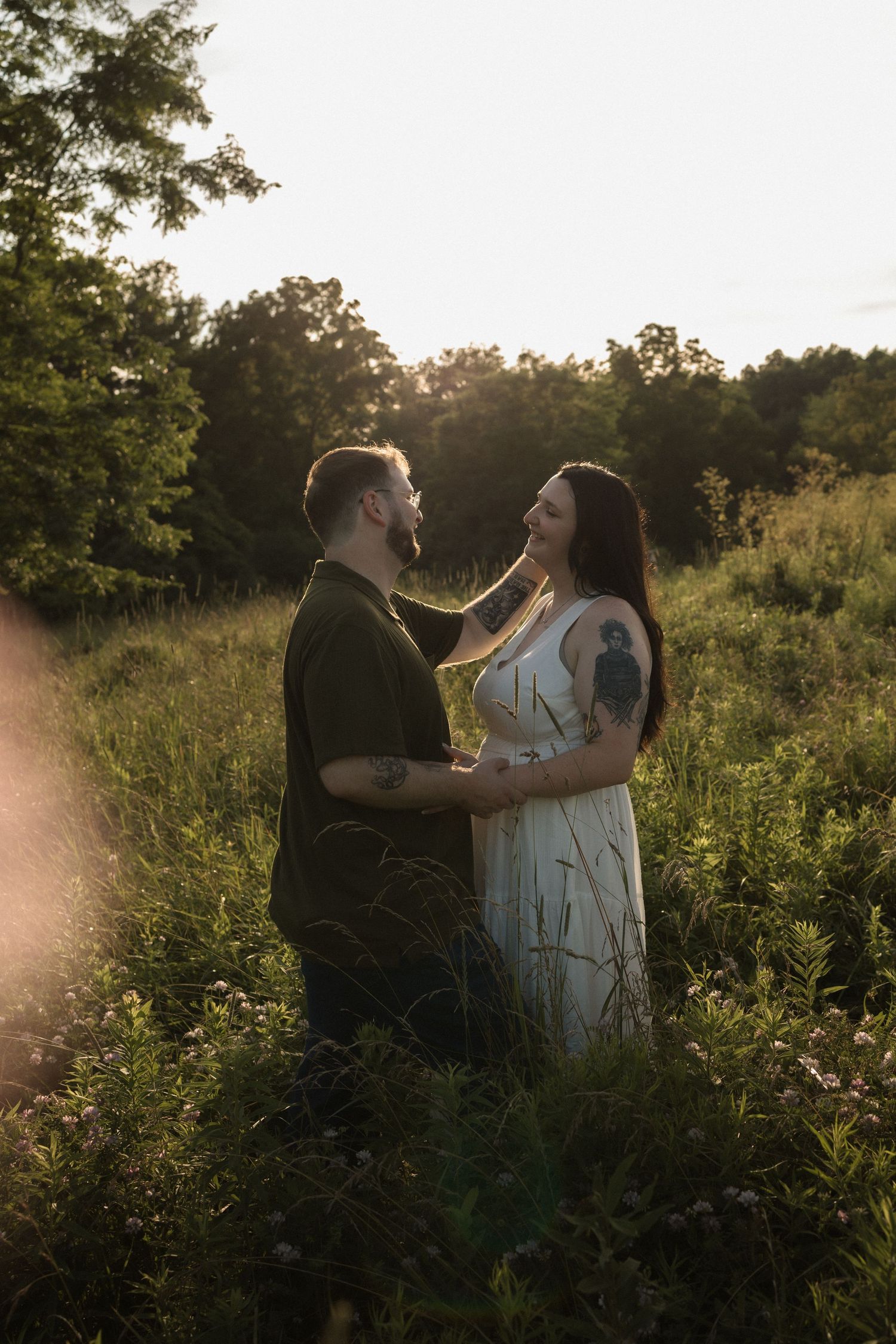 Couple embracing in a sunlit meadow at golden hour, woman in white dress touching mans face, surrounded by lush greenery.