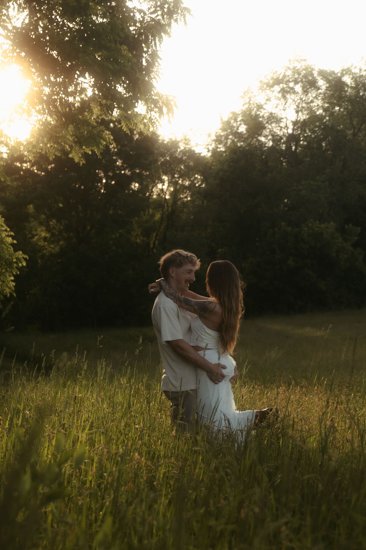 Couple embracing in golden sunset light in a lush green meadow surrounded by trees.