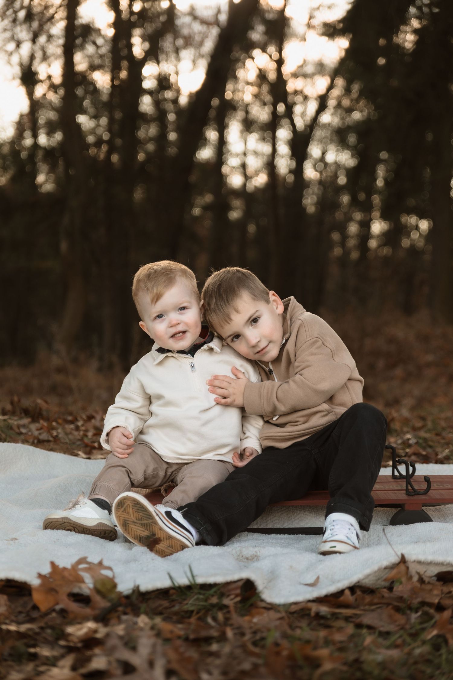 Two children sitting together on a white blanket surrounded by autumn leaves in a wooded setting.