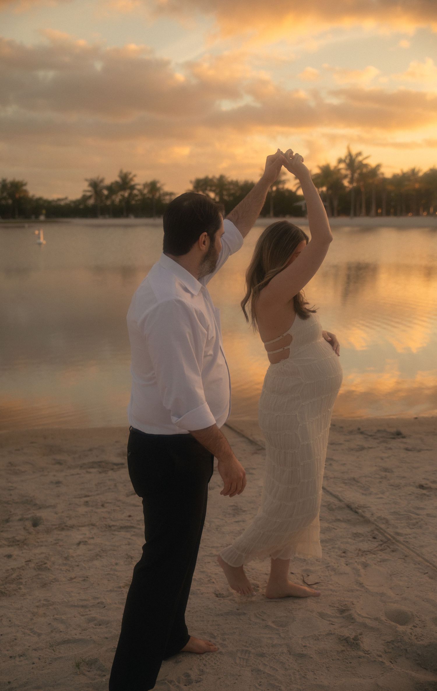 A couple dances together on a sandy beach at sunset with palm trees and calm waters in the background.