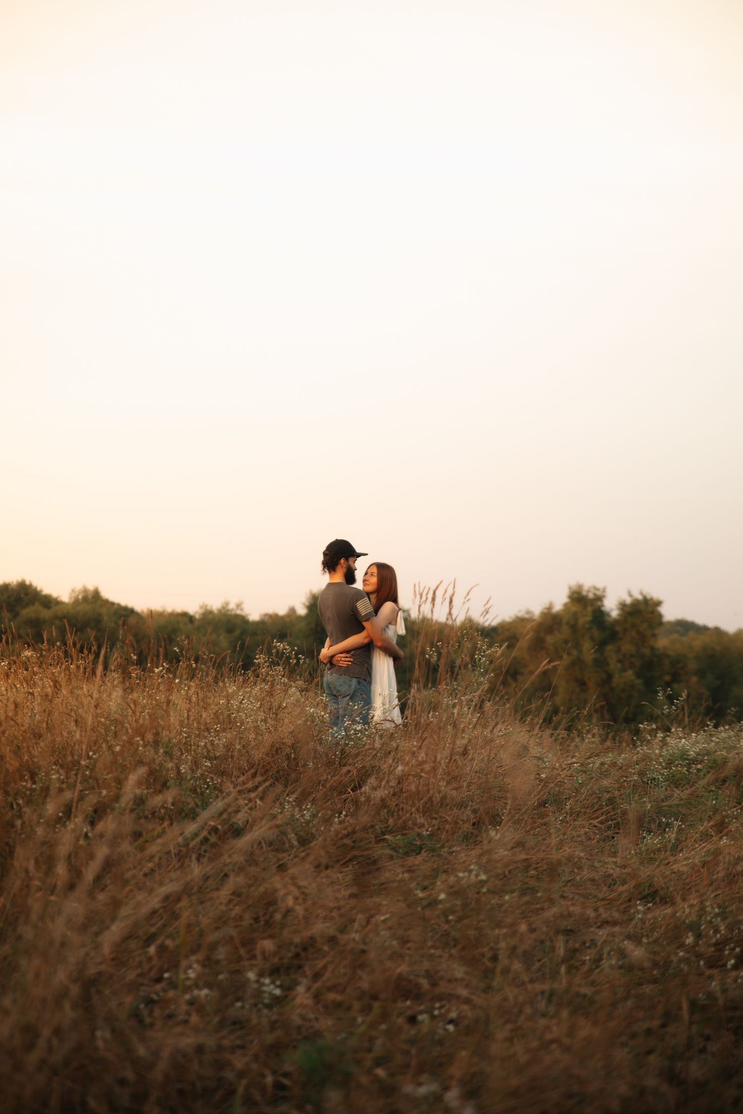 A couple embraces in a golden field at sunset, surrounded by tall dried grass and a warm glowing sky.