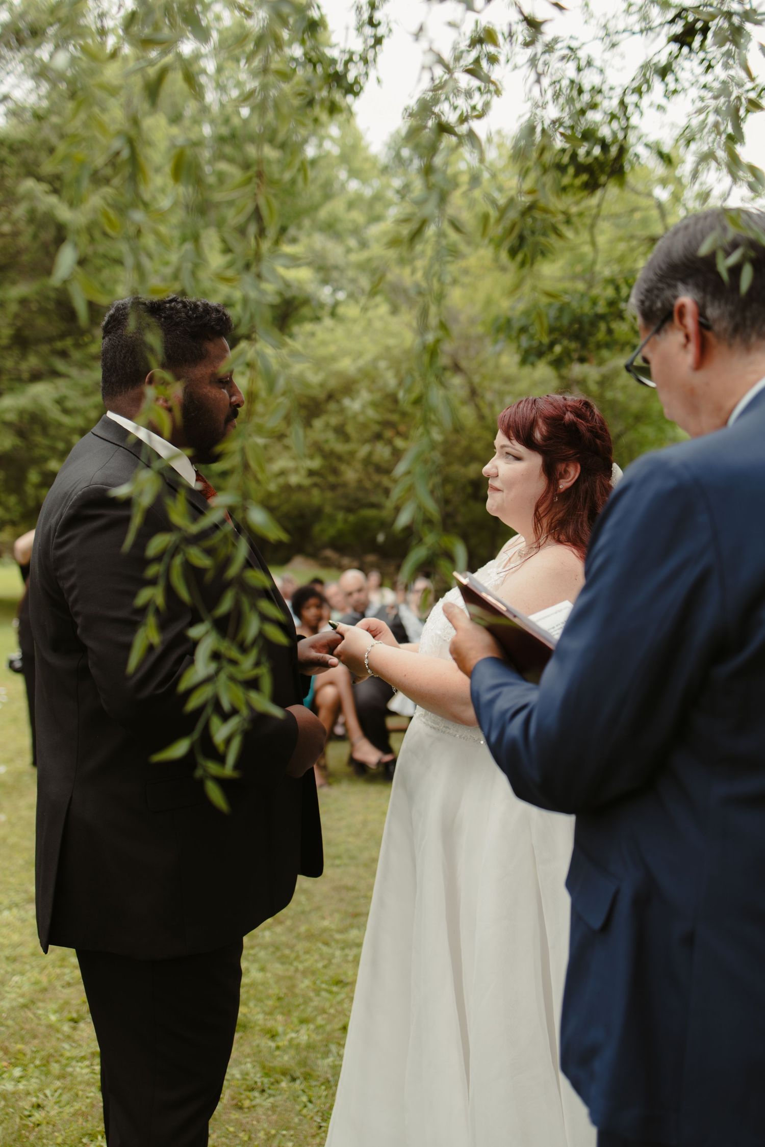 Outdoor wedding ceremony with bride in white dress and groom in black suit exchanging vows under willow tree.