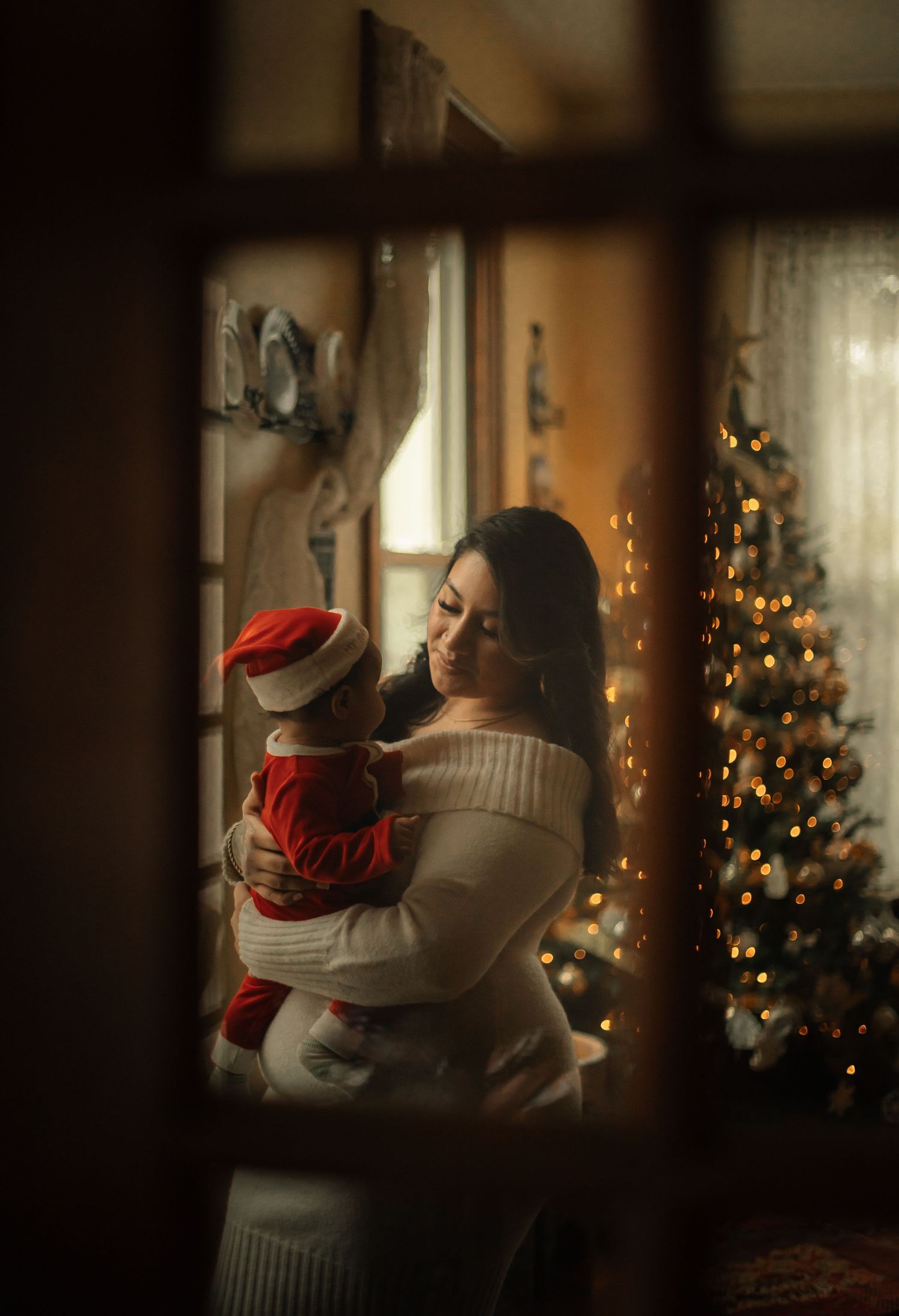 A mother in a white sweater holds her baby in a Santa hat near a glowing Christmas tree, viewed through a window.