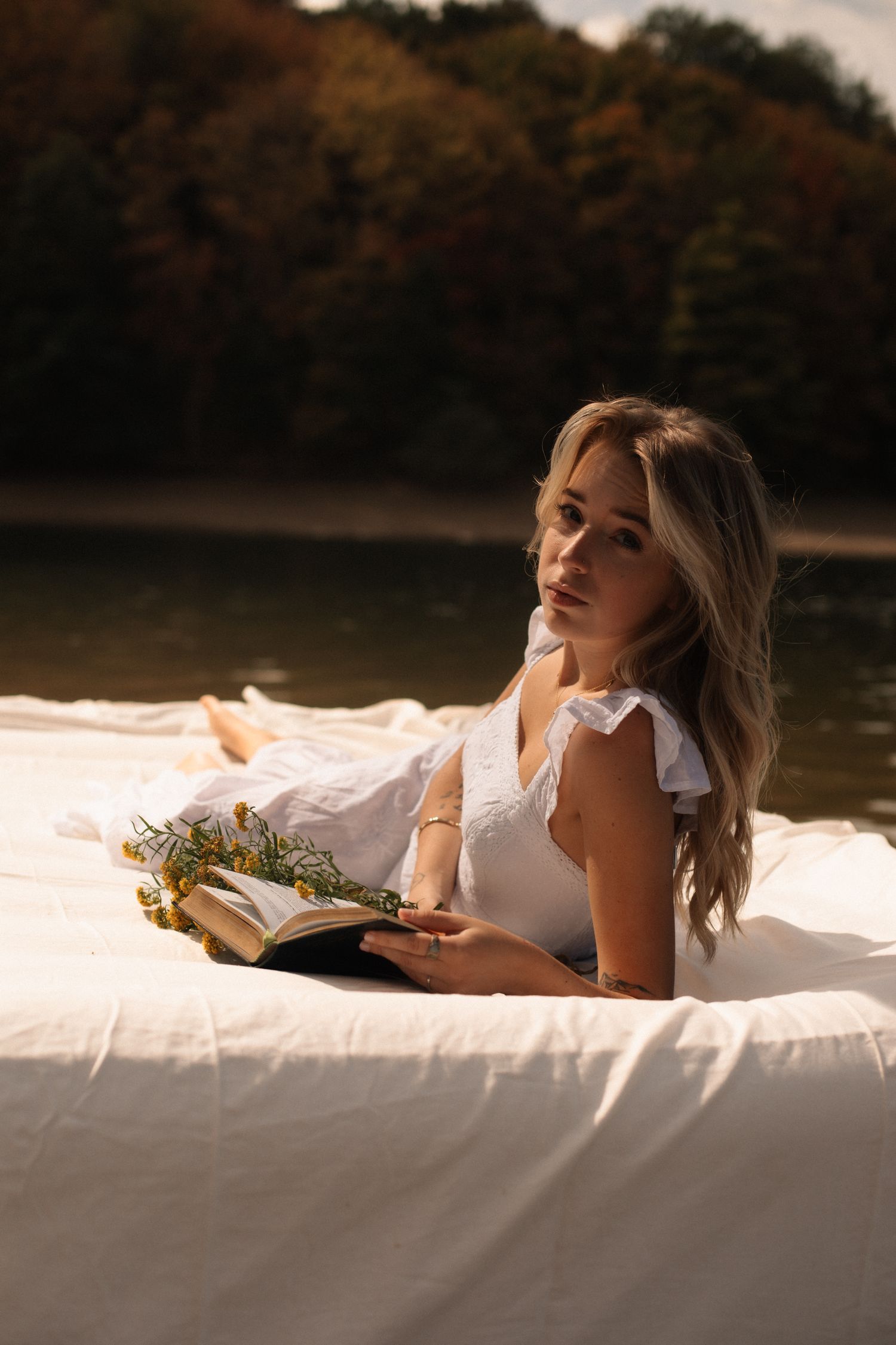 Young blonde woman in white dress lying on white blanket by lake, reading book with wildflowers, autumn trees background.