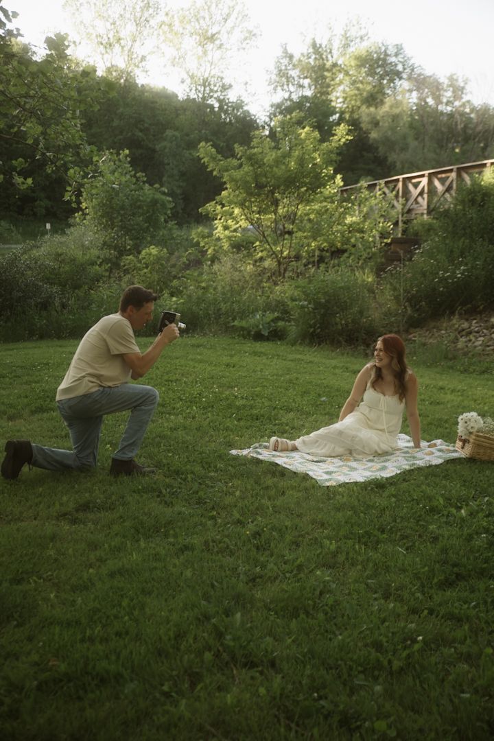 Man kneeling to photograph a woman in a white dress sitting on a blanket in a lush green park near a wooden bridge.
