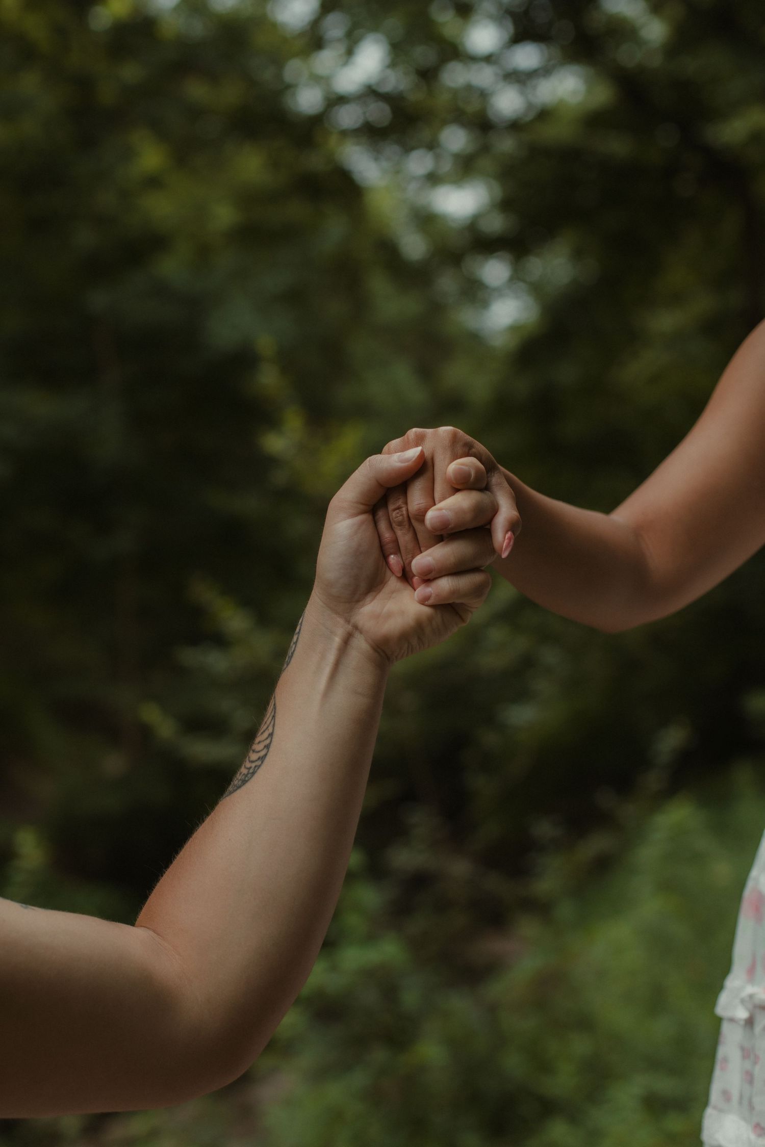Two people holding hands tenderly outdoors with lush green trees in the background, one hand showing a small tattoo.