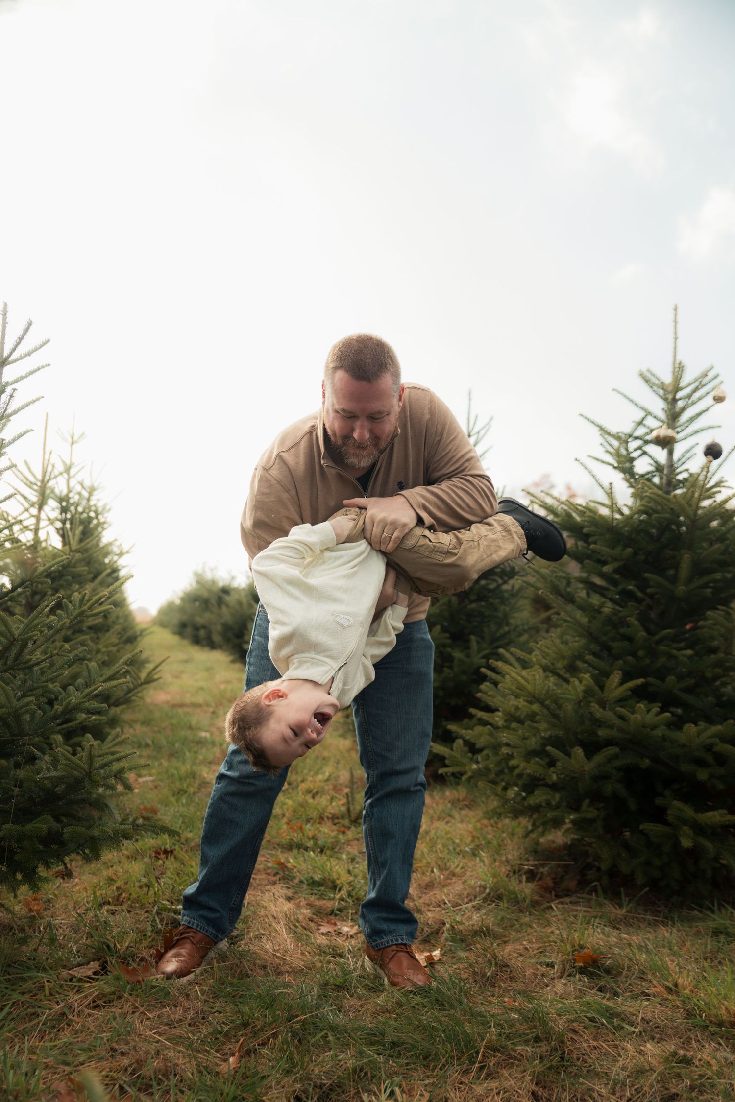 Two people playfully interact in a Christmas tree farm surrounded by evergreen trees on a cloudy day.