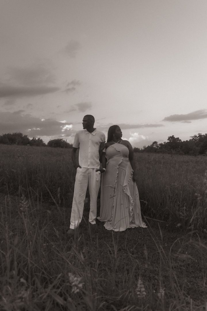 Black and white photo of a couple standing together in a field at sunset, viewed from behind.